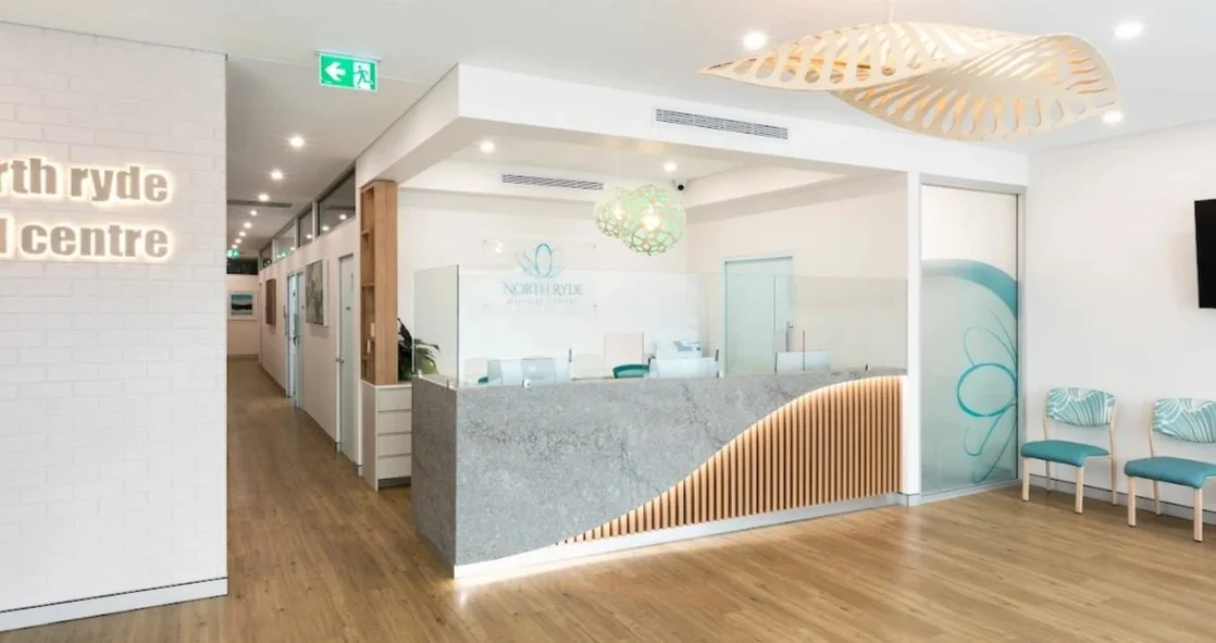 Reception area of North Ryde Dental Practice with a modern gray marble front desk, teal chairs, and decorative light fixtures, with a hallway leading to private rooms.