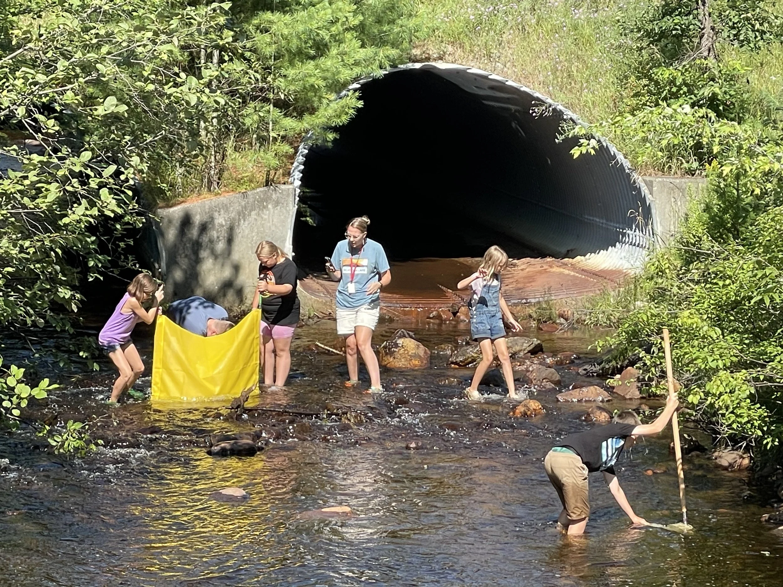 Group of children and adults in a creek near a large pipeline tunnel, some holding a yellow fabric, one person with a stick, surrounded by trees and bushes.