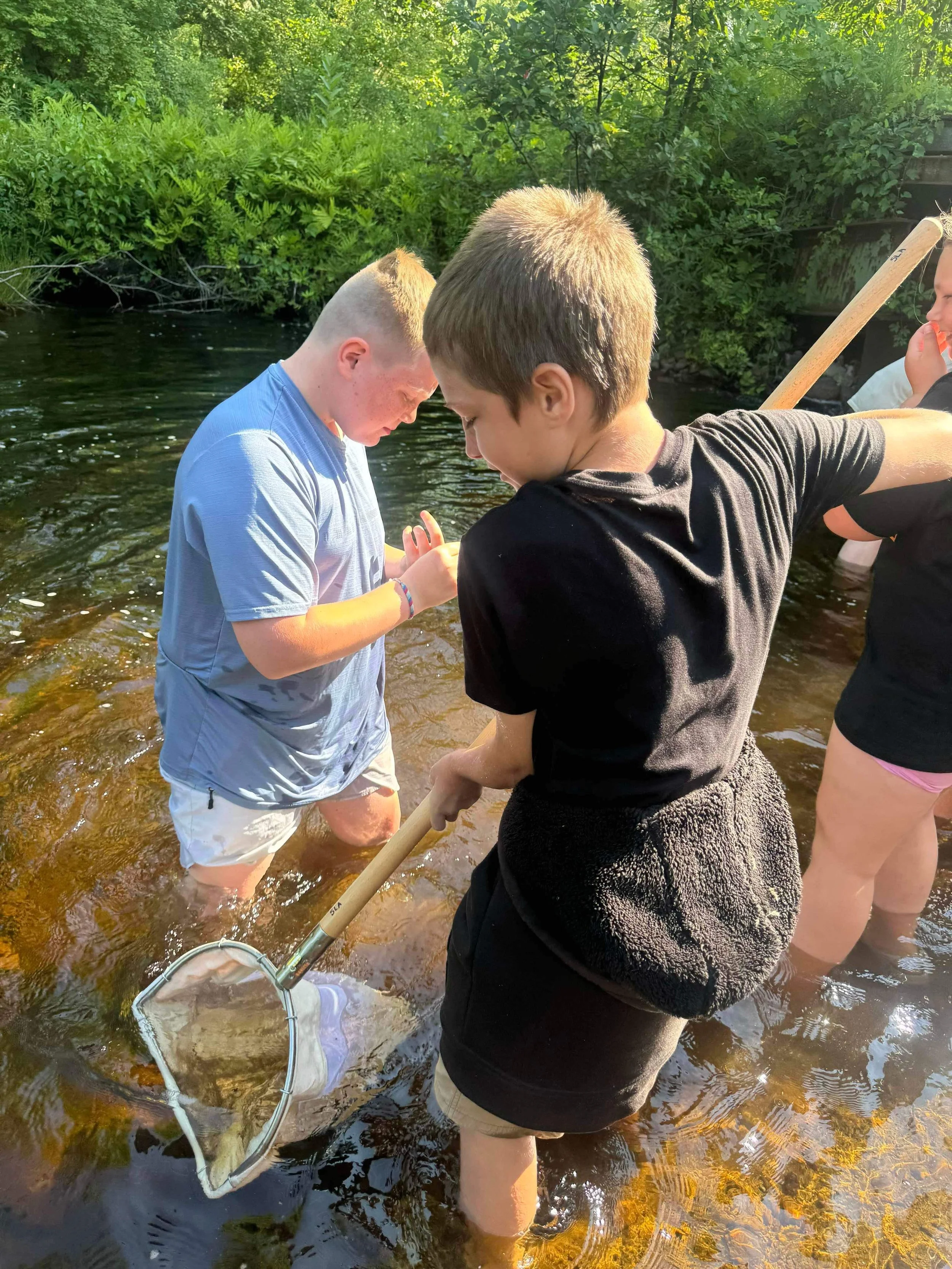 Three children wading in a shallow river surrounded by greenery, one holding a fishing net while the other two look at something in their hands.