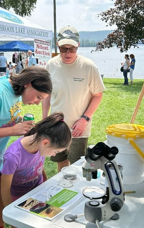 A woman and two girls examine something through a microscope at an outdoor event by a lake, with tents and other people in the background.