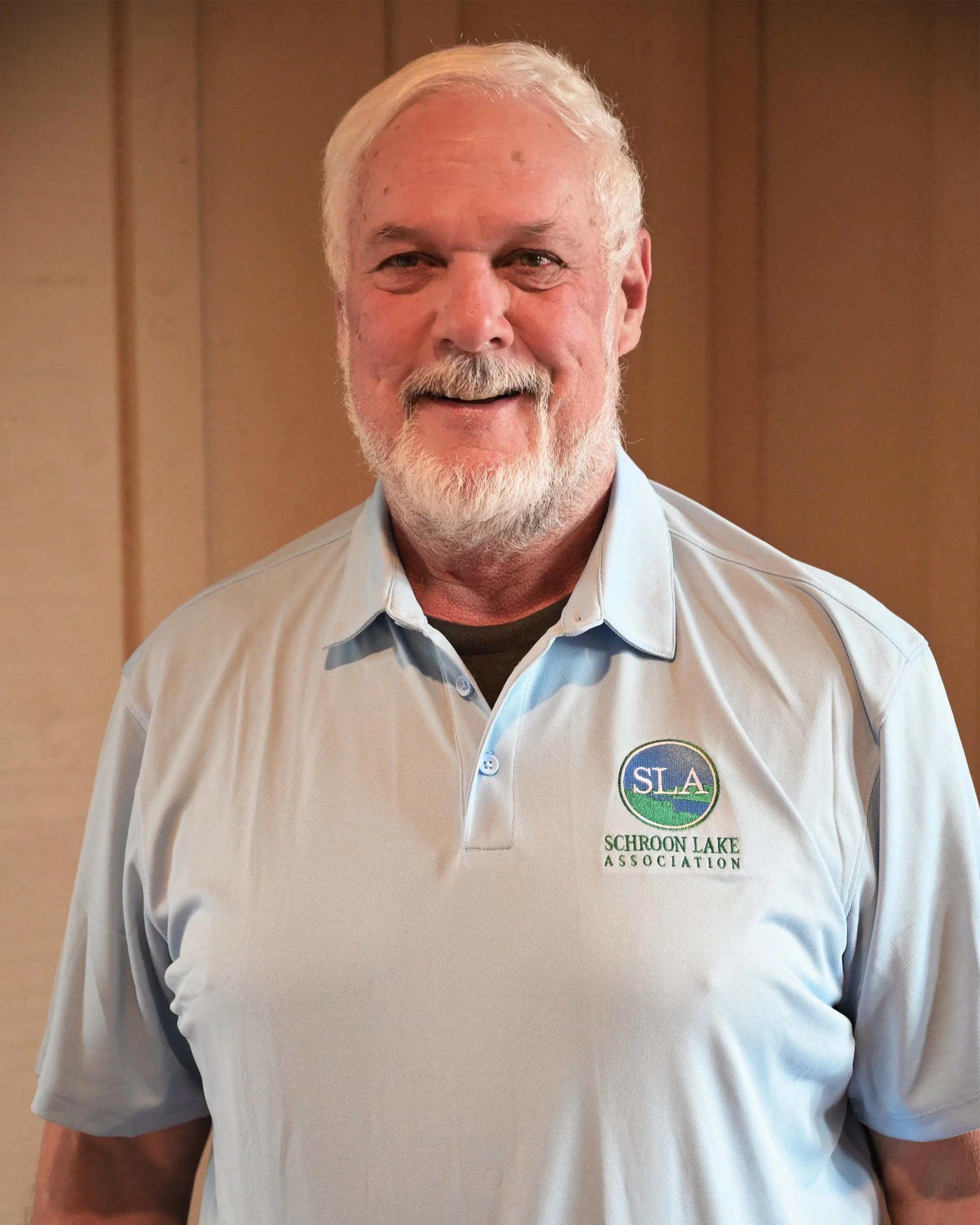 A smiling elderly man with white hair and a beard wearing a light grey polo shirt with a logo that says 'SLA Schroon Lake Association' on the left side.