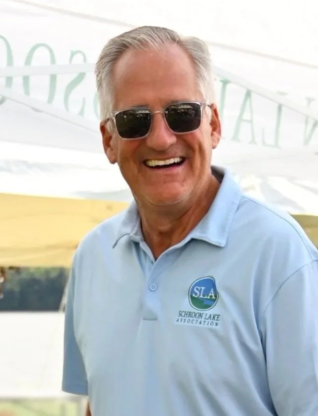 A smiling man wearing sunglasses and a light blue polo shirt with a logo that says 'SLA Schulson Lake Association'.