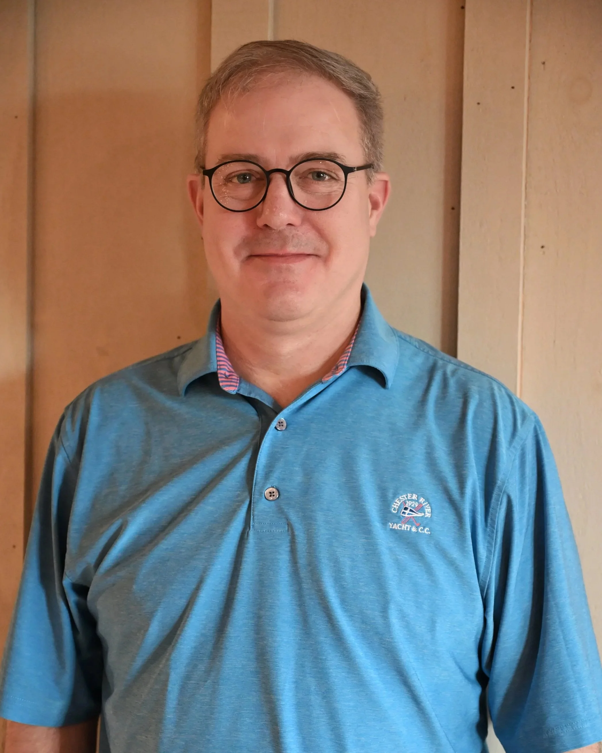 Man wearing glasses and a blue polo shirt with a logo, standing indoors against wooden paneling.