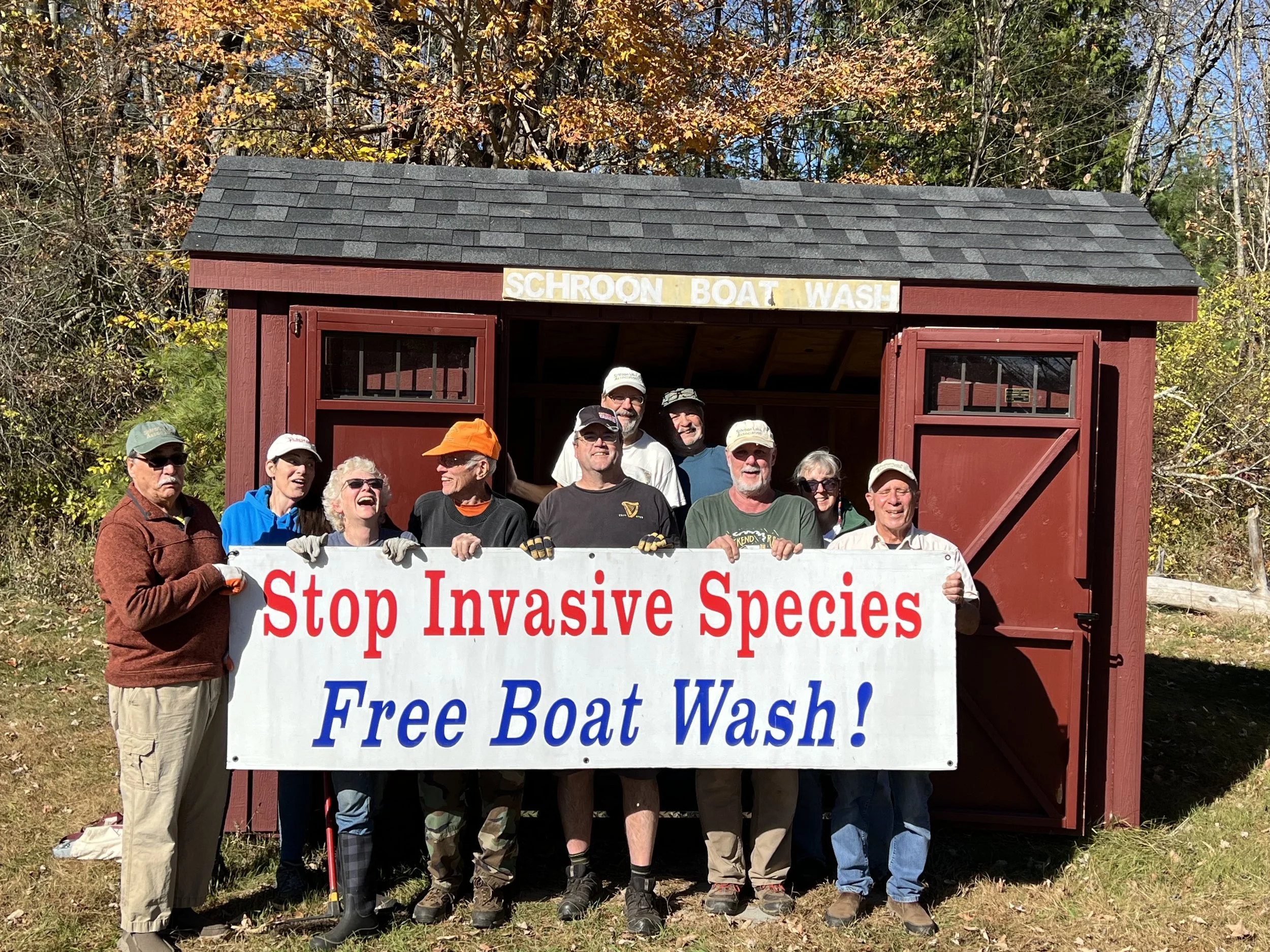 Group of people holding a large sign that reads 'Stop Invasive Species Free Boat Wash!' in front of a small red wooden shed labeled 'Schoon Boat Wash,' surrounded by trees with fall foliage.