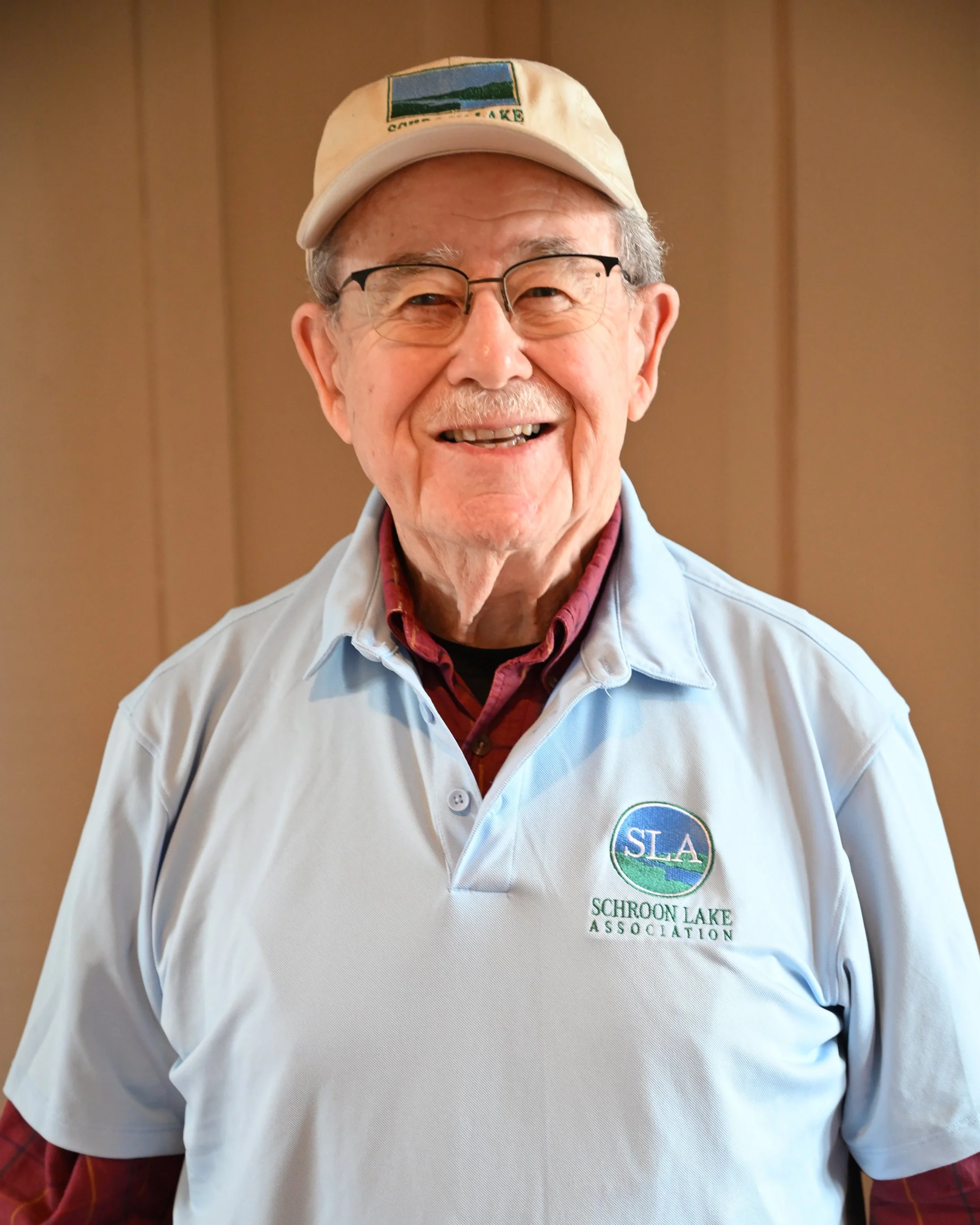 An elderly man with glasses and a smile, wearing a beige cap and a white polo shirt with a logo that says 'SLA Schroon Lake Association', standing in front of a wooden wall.