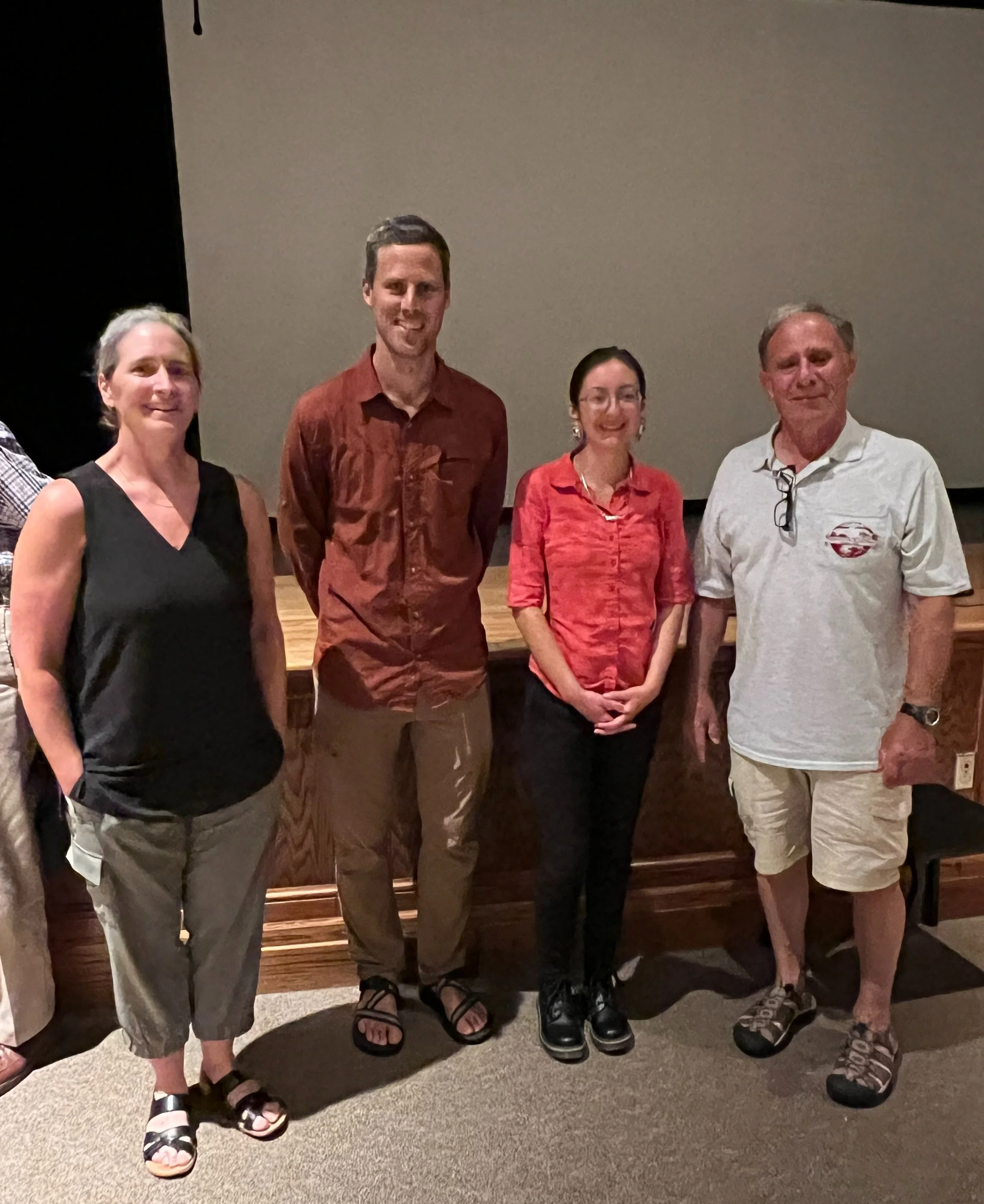 Group of four people standing together on a stage in front of a blank wall, smiling at the camera.