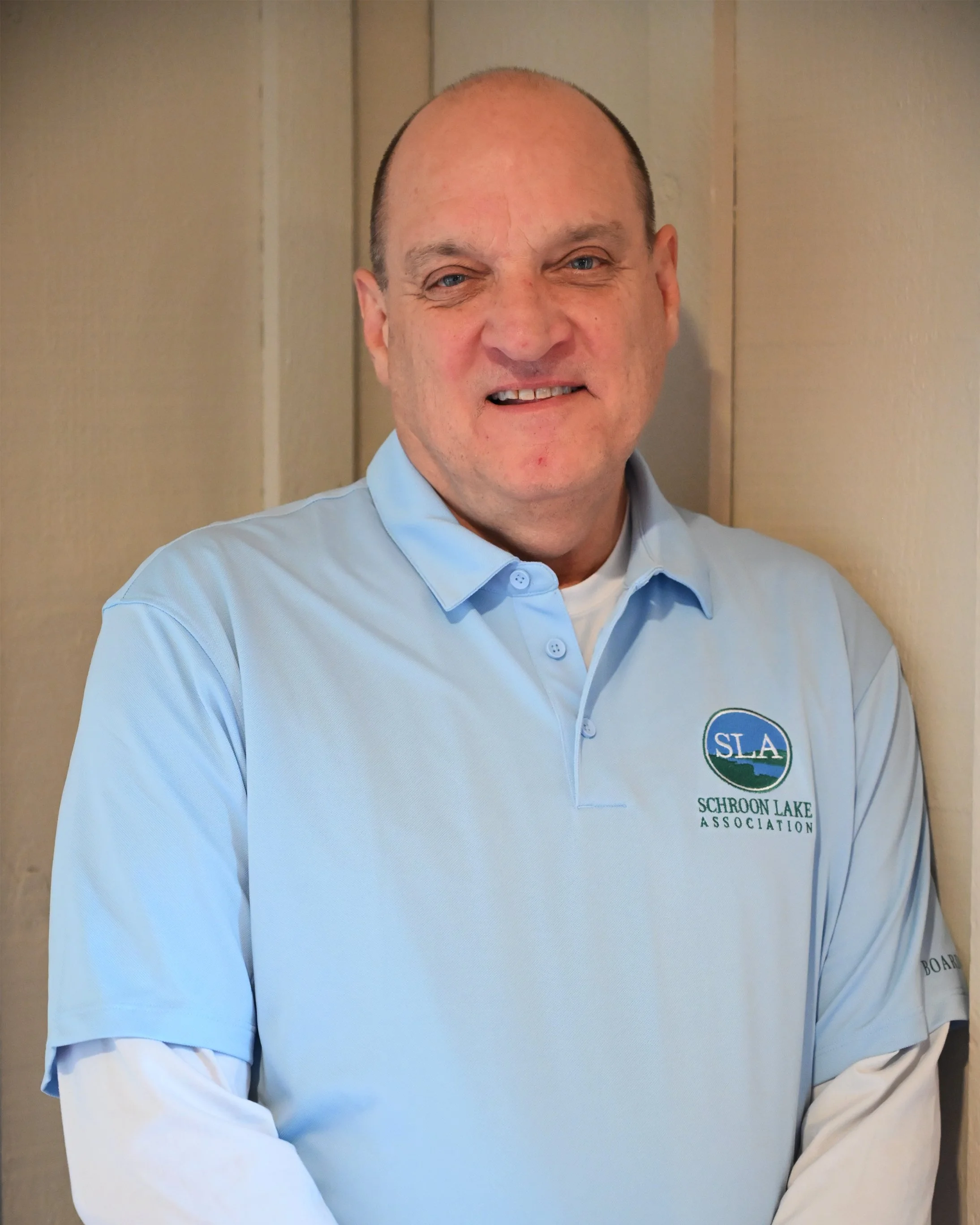 A man with a shaved head wearing a light blue polo shirt with a logo of Schroon Lake Association stands against a beige wall and smiles.