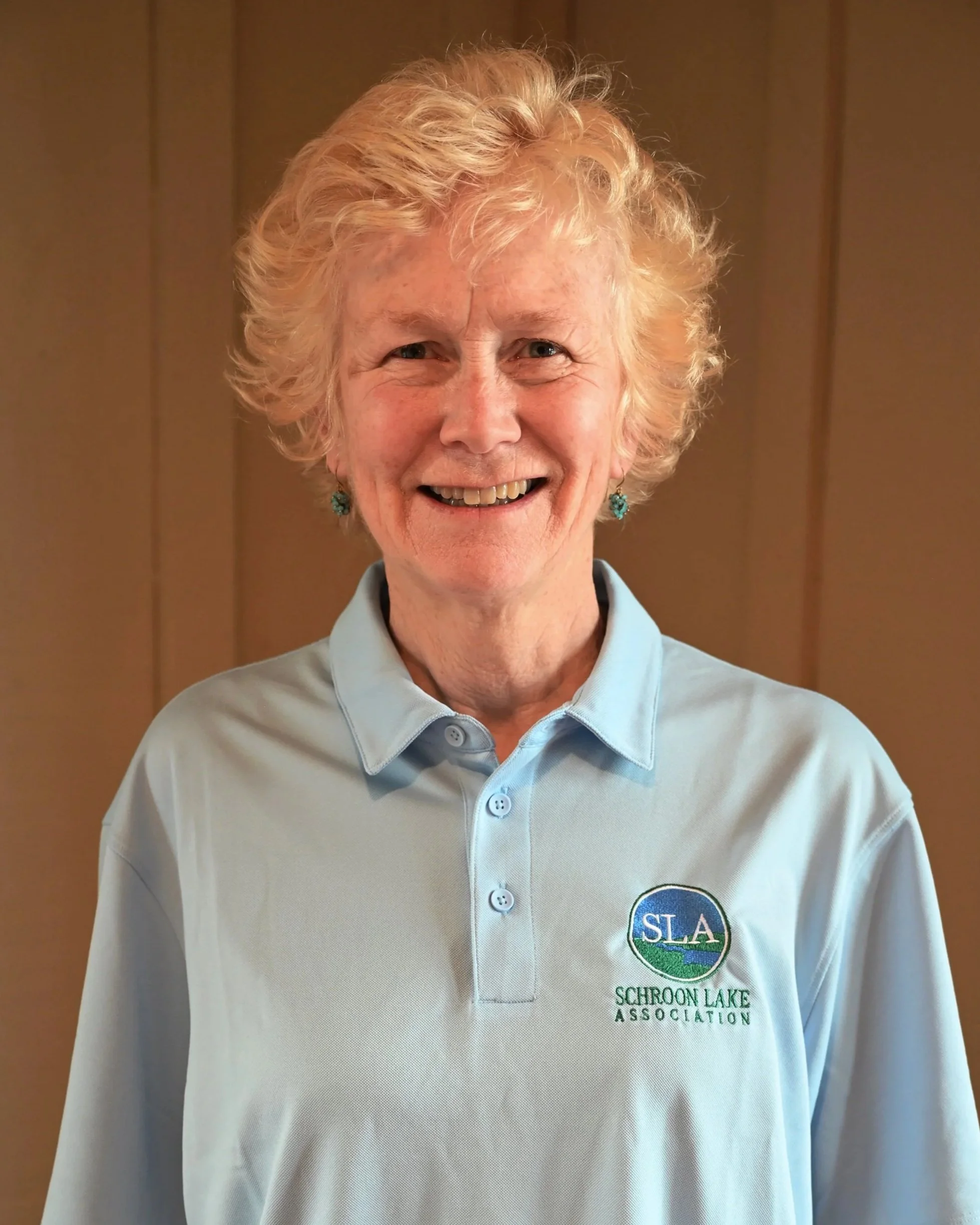 A smiling elderly woman with curly blonde hair, wearing a light blue polo shirt with the logo of Schrooon Lake Association.