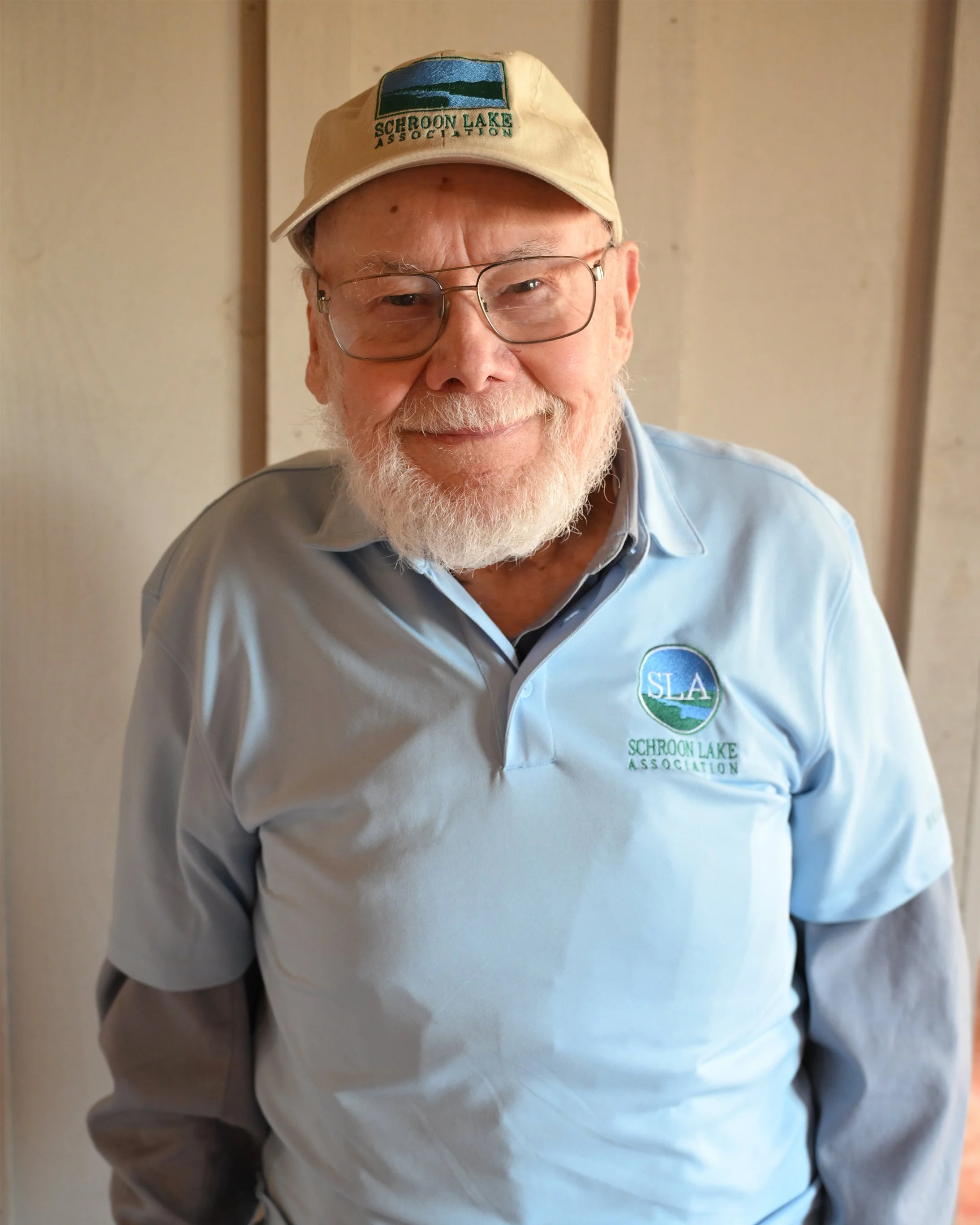 A smiling elderly man with glasses and a white beard wearing a beige cap with a 'Schroon Lake Association' logo and a light blue polo shirt with the same logo.