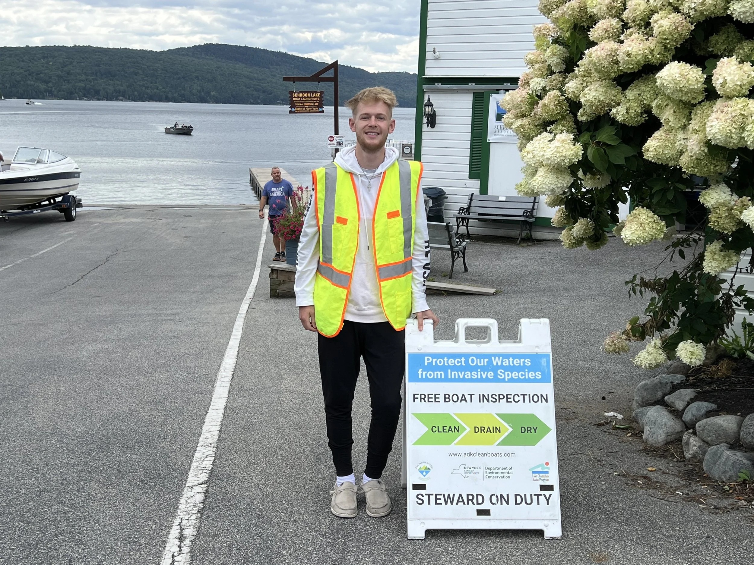 Young man in a high-visibility safety vest standing next to a sign that promotes boat waterway protection and offers free boat inspections, with a lake and boat in the background.