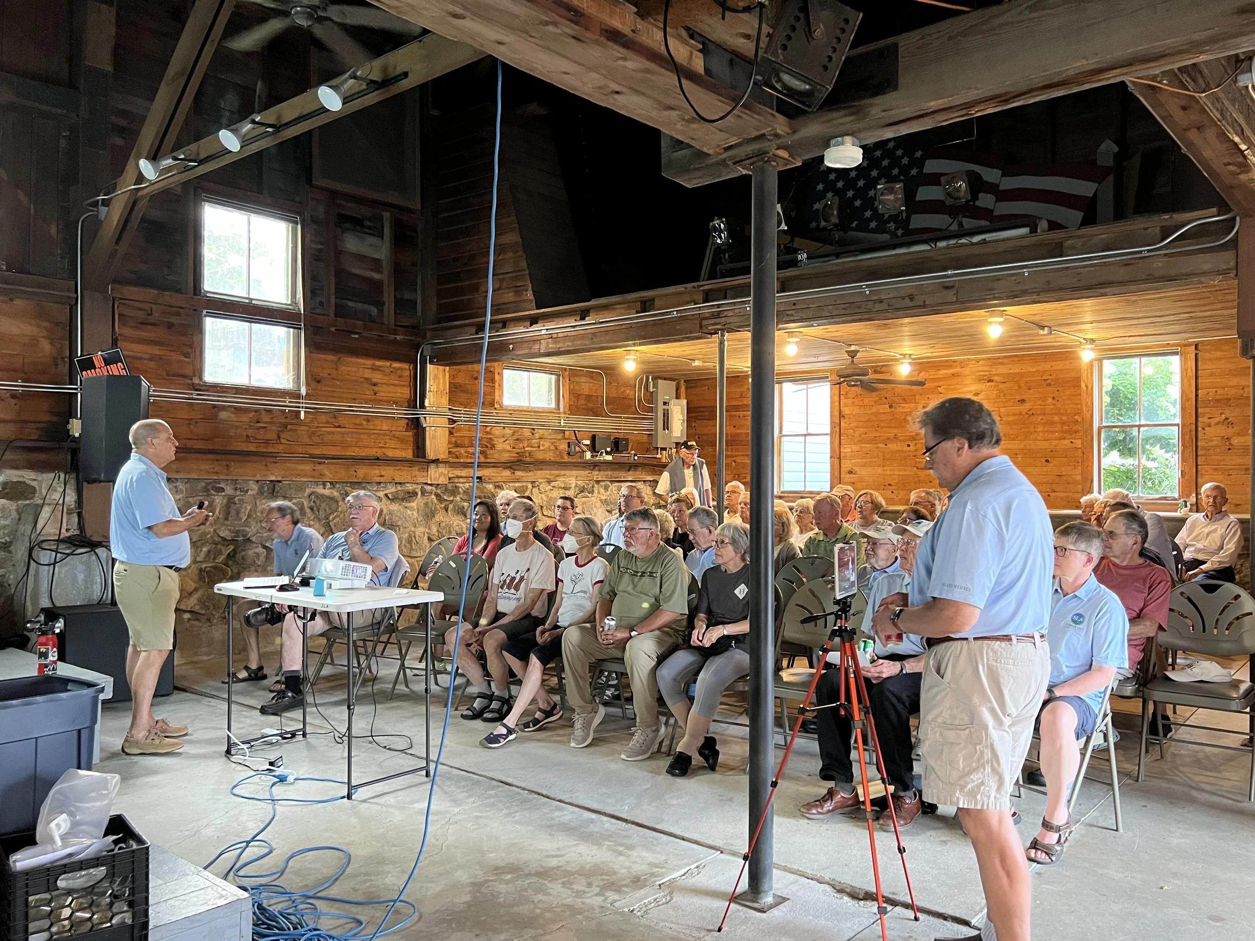 Man in blue shirt and khaki shorts giving a presentation to an audience in a rustic wooden hall with stone wall accents. The audience, mainly older adults, are seated on plastic chairs, some wearing masks, and appear to be attentively listening. There is a projector on a table and a person operating a camera on a tripod.