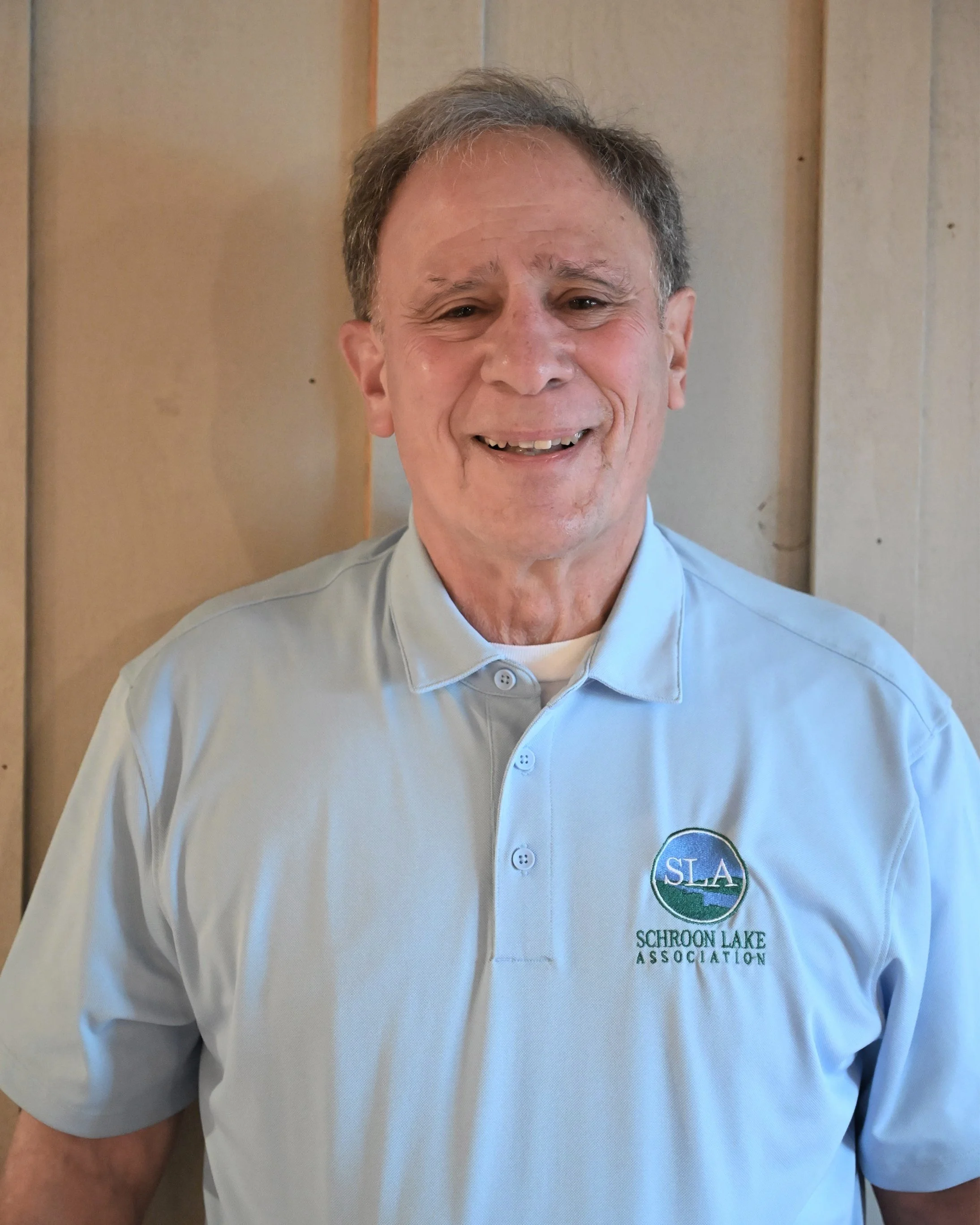 Smiling middle-aged man in light blue polo shirt with 'Schroon Lake Association' logo, standing in front of wood-paneled wall.