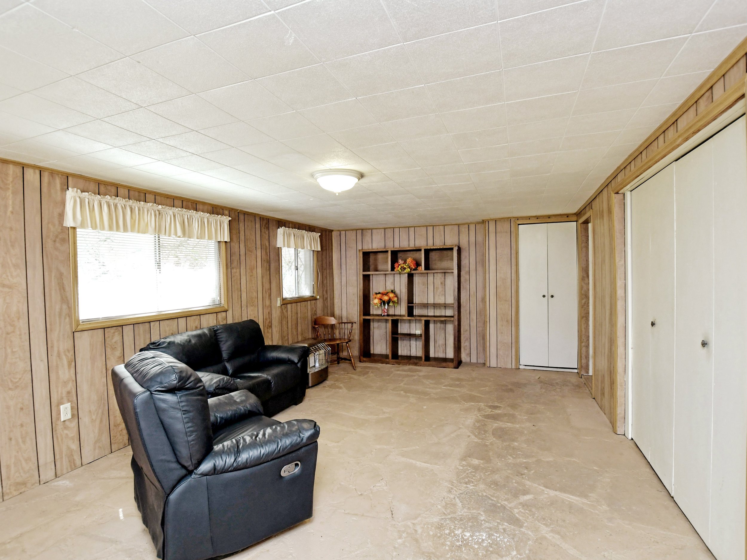 A basement with wood paneling, featuring a black leather recliner and sofa, a wooden bookshelf with decorative items, a small side table, windows with curtains, and a door leading to another room.