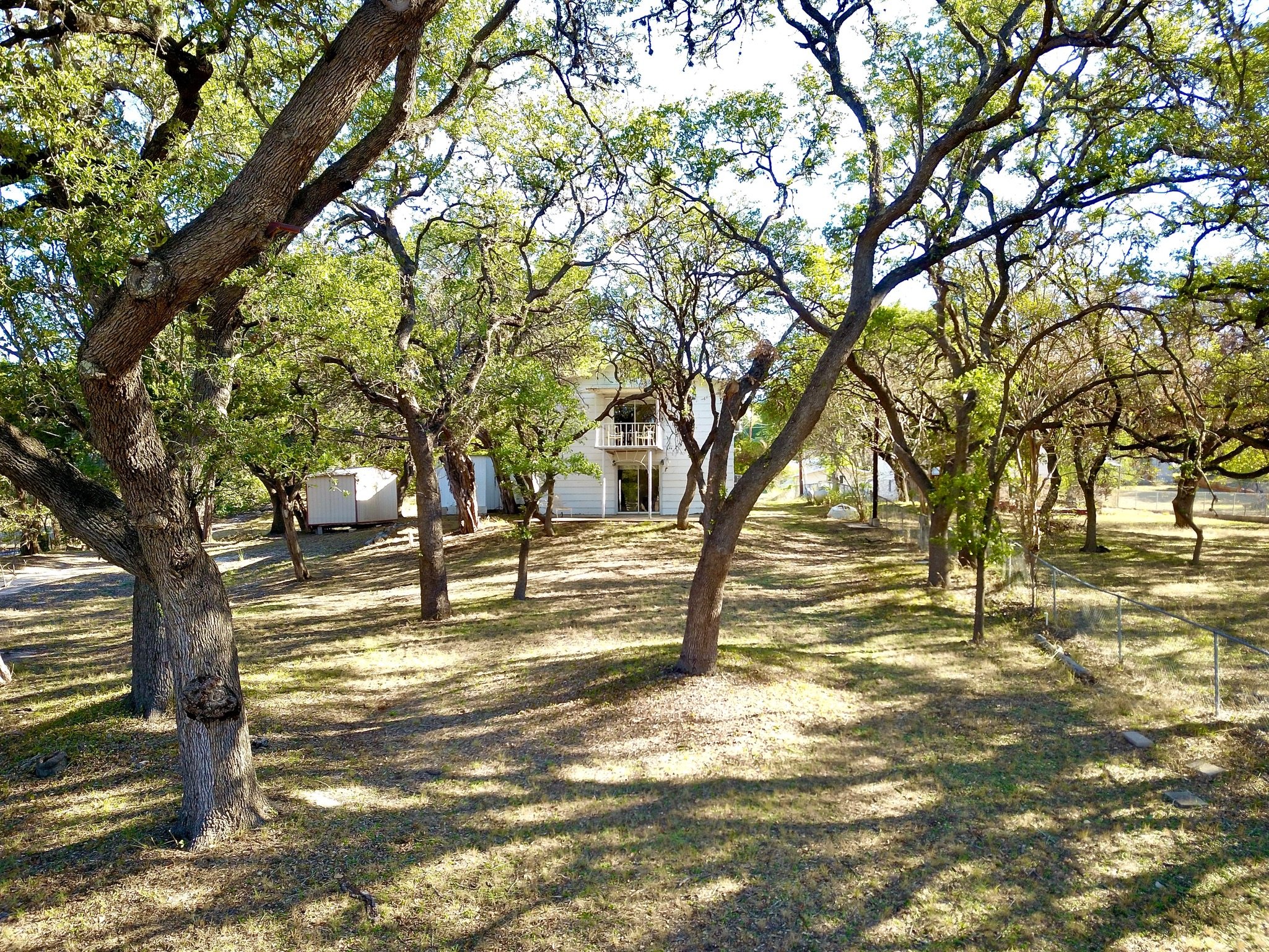 A white house surrounded by trees in a wooded area, with a small shed nearby and a wire fence along the right side.