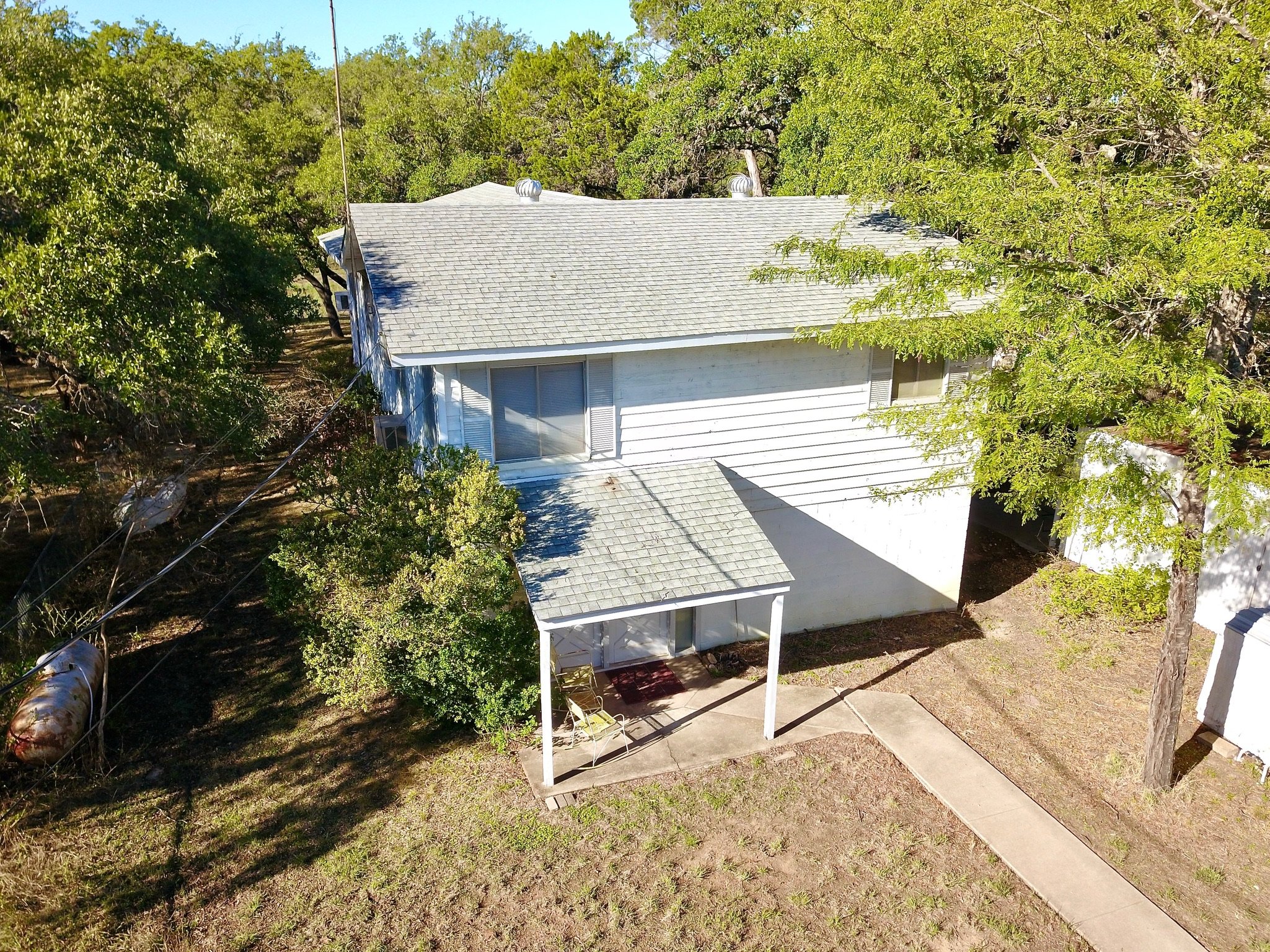 Aerial view of a two-story house with a light gray roof, surrounded by trees and greenery. The house has a small porch and a paved walkway leading to it, set in a rural area.