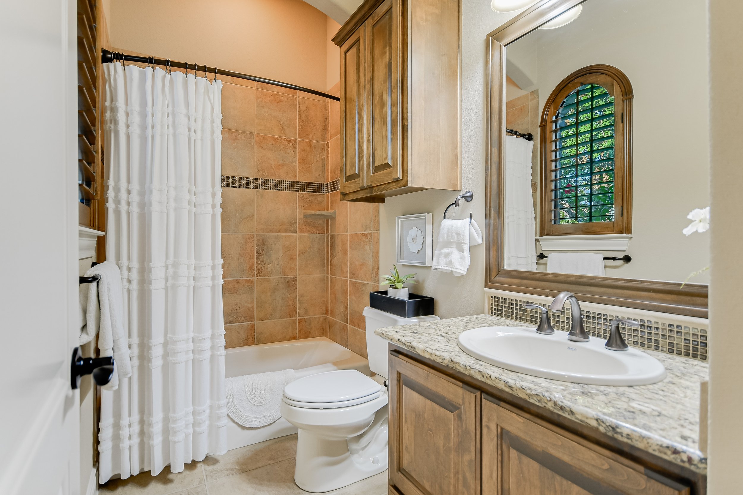 Modern bathroom with granite countertop, white sink, wooden cabinets, tiled shower, white curtain, toilet, and arched window.