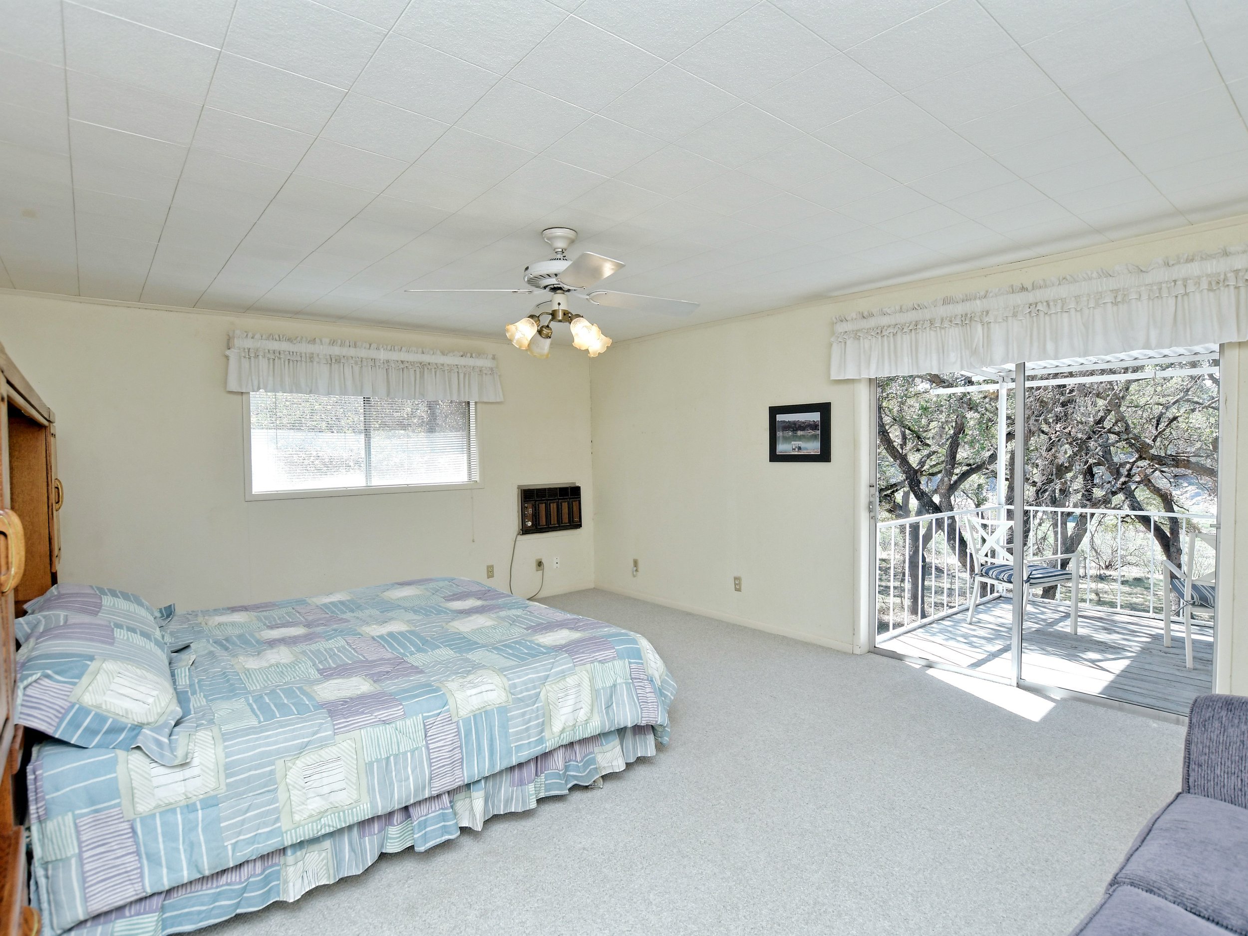 Spacious bedroom with large bed, ceiling fan, and sliding glass doors leading to a balcony with outdoor seating overlooking trees.