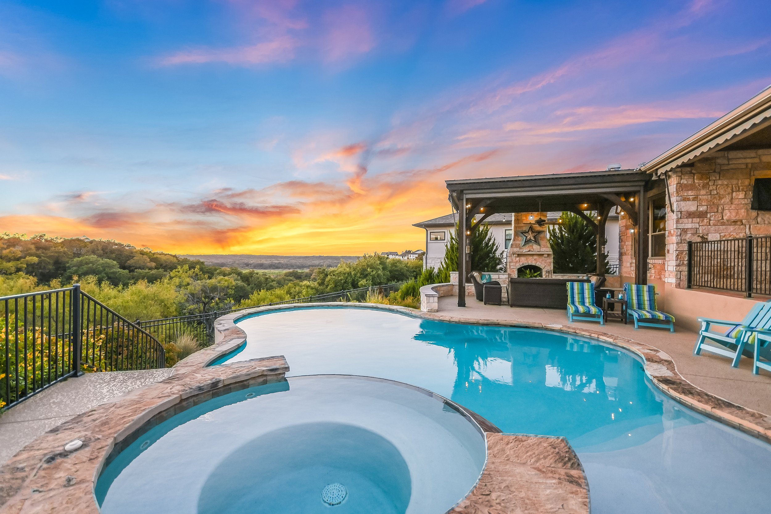Scenic backyard with swimming pool and sunset views, featuring a stone patio, pergola, outdoor seating, and lush greenery.