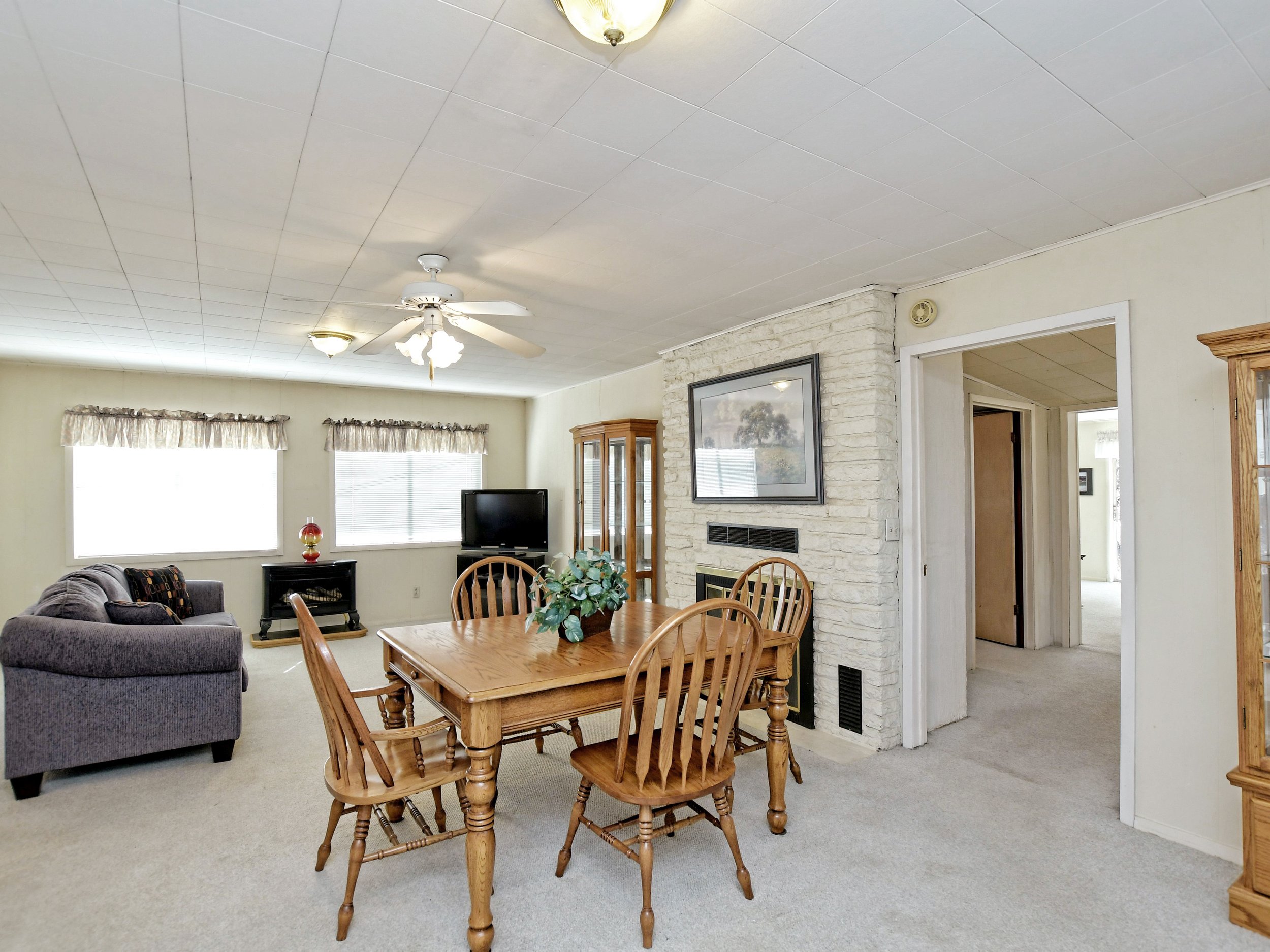 Open living and dining area in a home with a brick fireplace, wooden dining table with chairs, gray sofa, two large windows with curtains, ceiling fans, and a cabinet with a glass door.