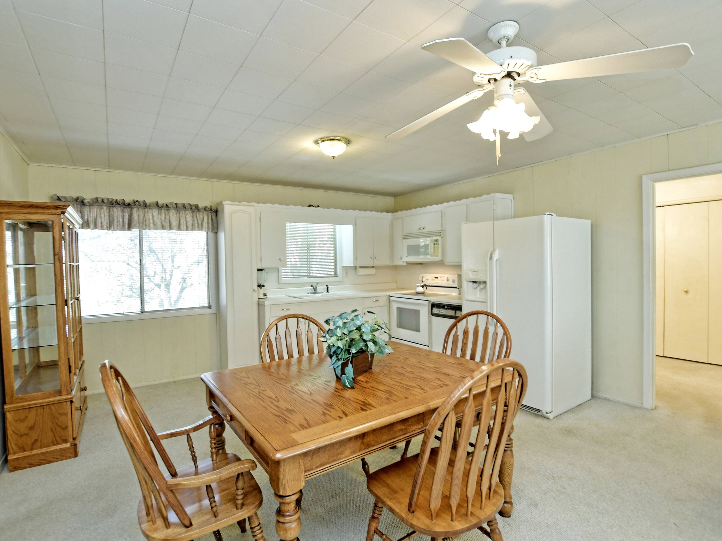 Kitchen and dining area with a wooden table and four chairs, white refrigerator, stove, microwave, cabinets, a ceiling fan with lights, a window with curtains, and a wooden display cabinet.