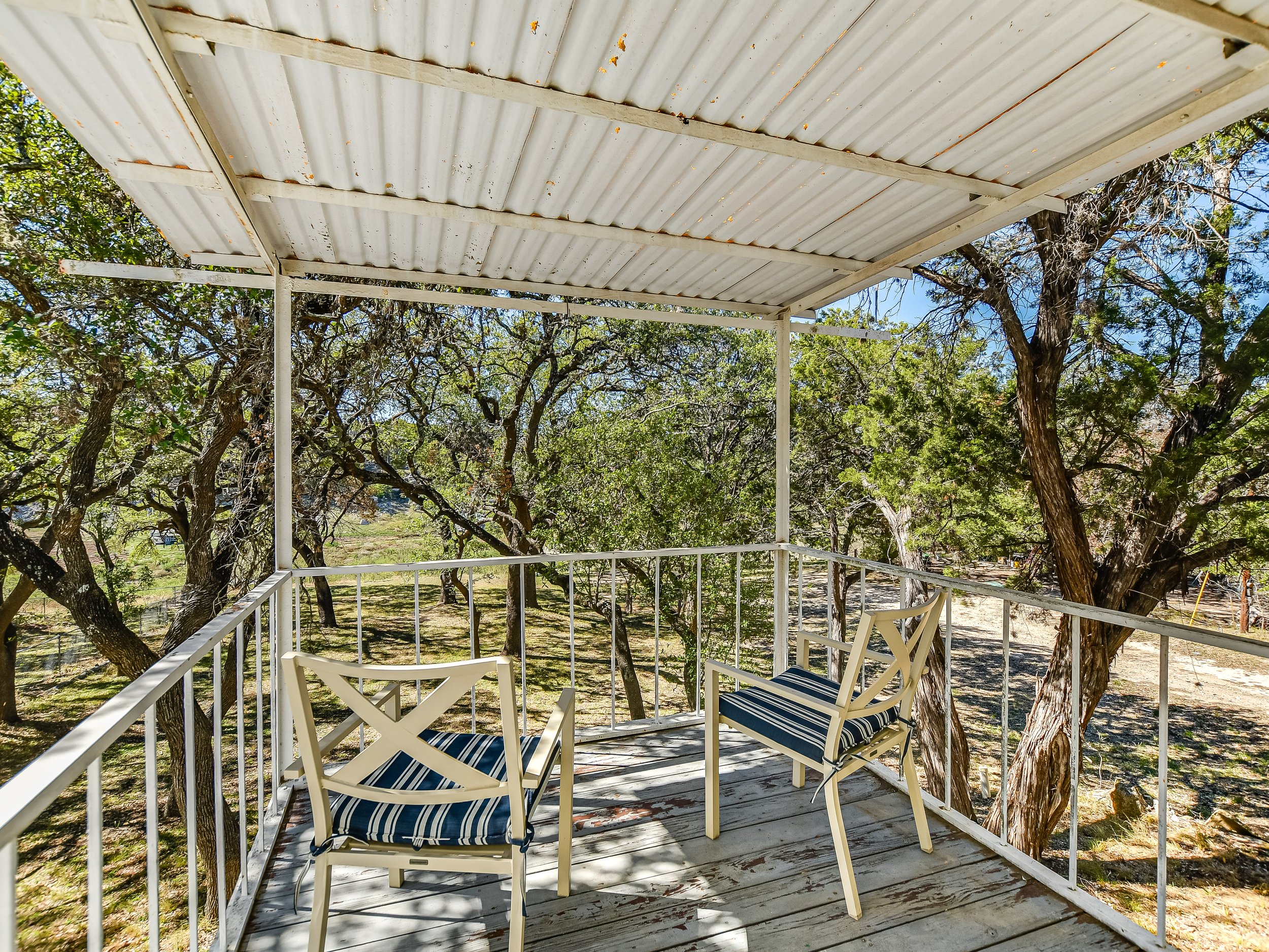 Outdoor porch with white metal roof, two cushioned chairs, surrounded by trees, overlooking a natural setting.