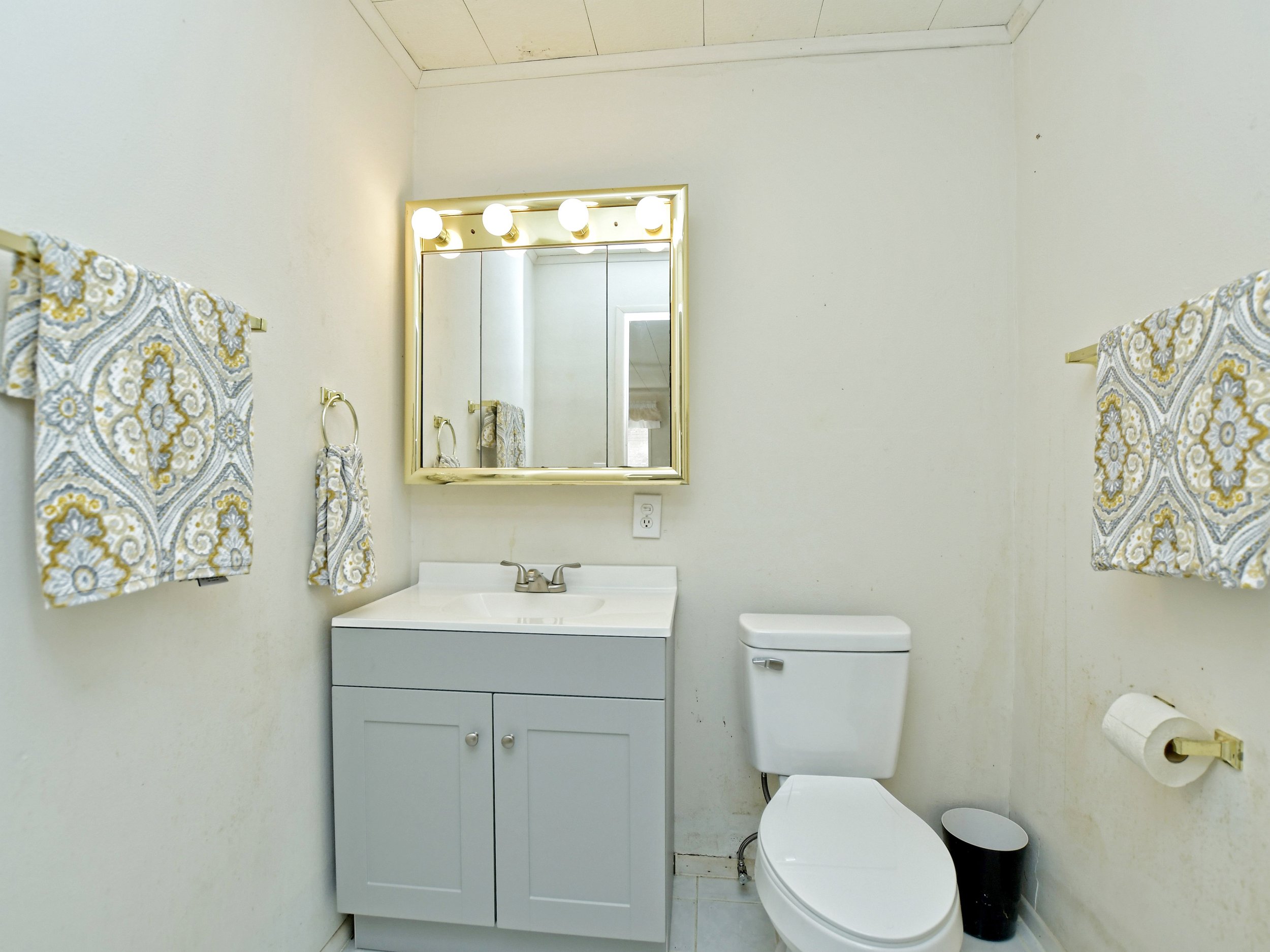 Small bathroom with a gray vanity, mirror with lights, toilet, patterned towels, toilet paper, and wastebasket.