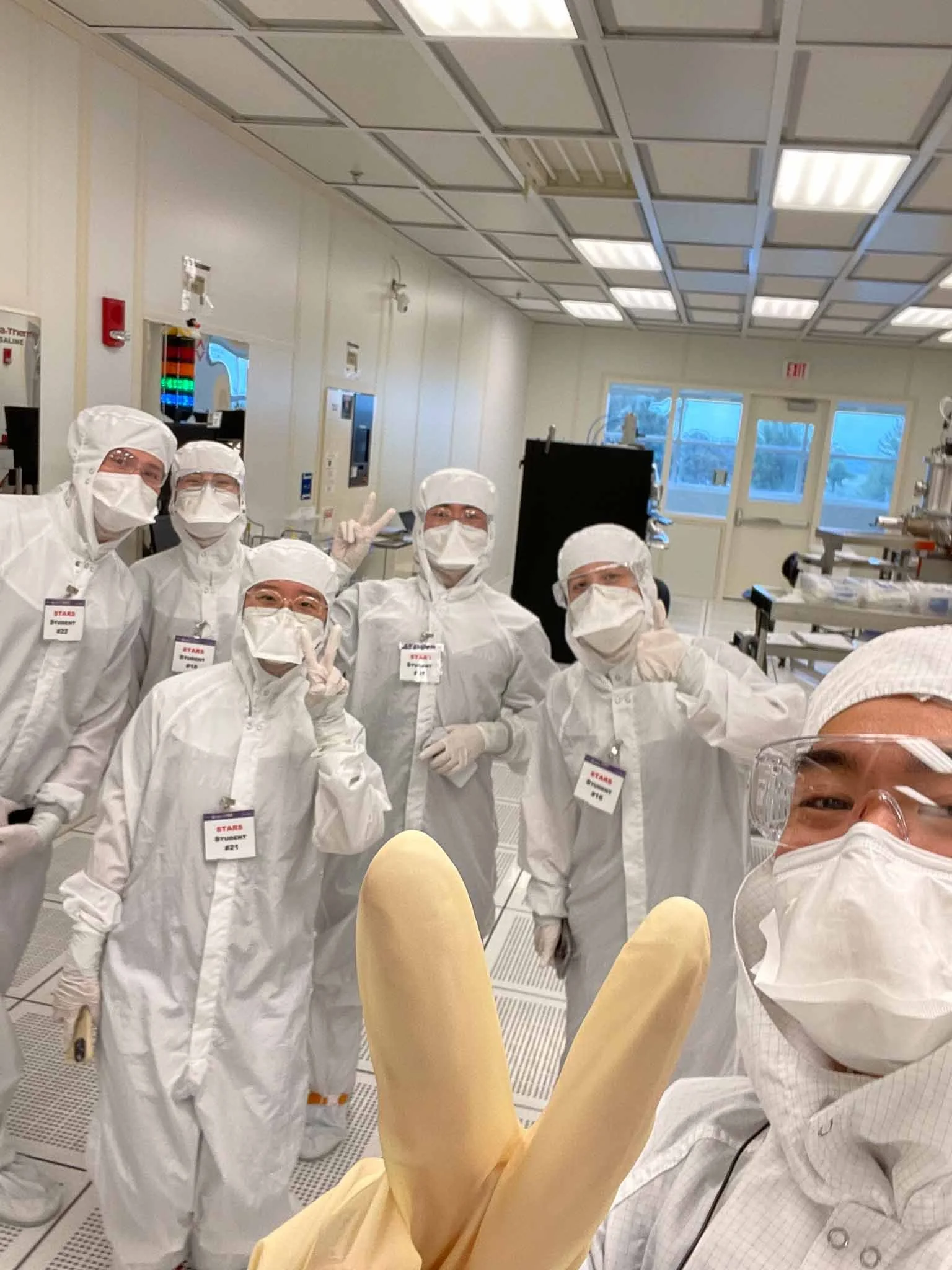 Group of scientists and researchers in a laboratory wearing white protective suits, masks, gloves, and goggles, posing for a group photo.