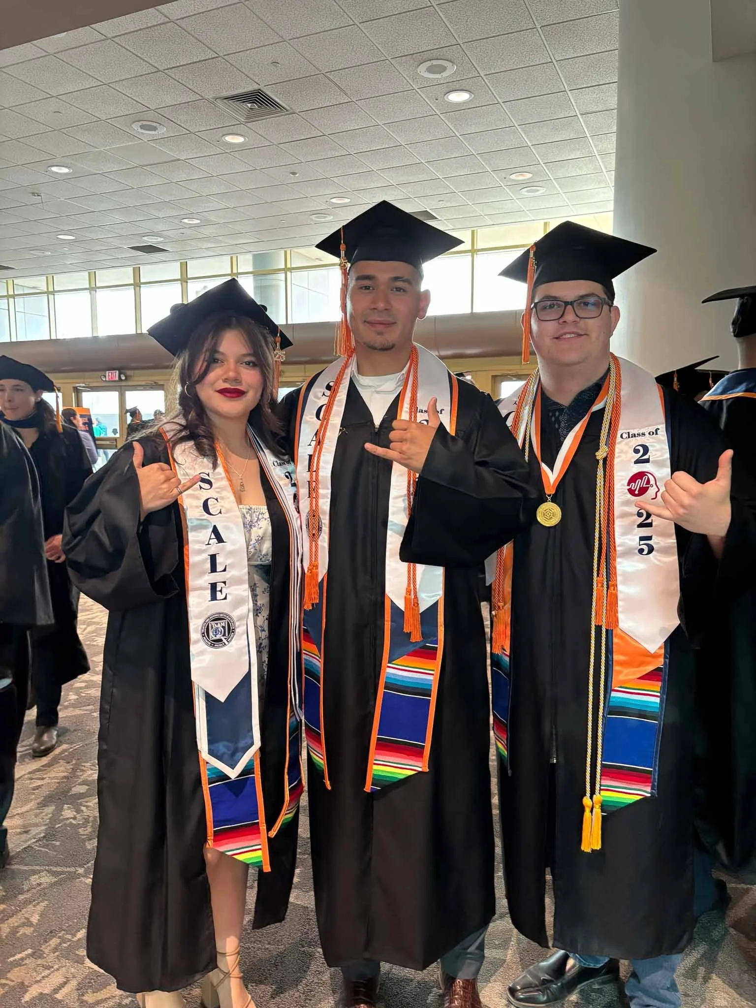 Three graduates in caps and gowns at a graduation ceremony, making hand gestures, with other graduates in the background.
