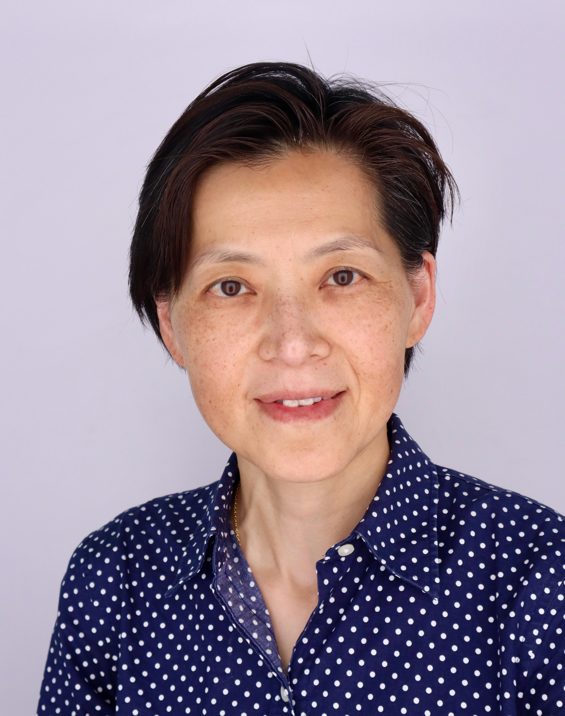 Professional portrait of a woman with short black hair, wearing a dark blazer and a light blue collared shirt, smiling against a gray background.