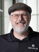 A man with glasses, a beard, and a mustache wearing a dark flat cap and a navy blue polo shirt, smiling against a plain background.