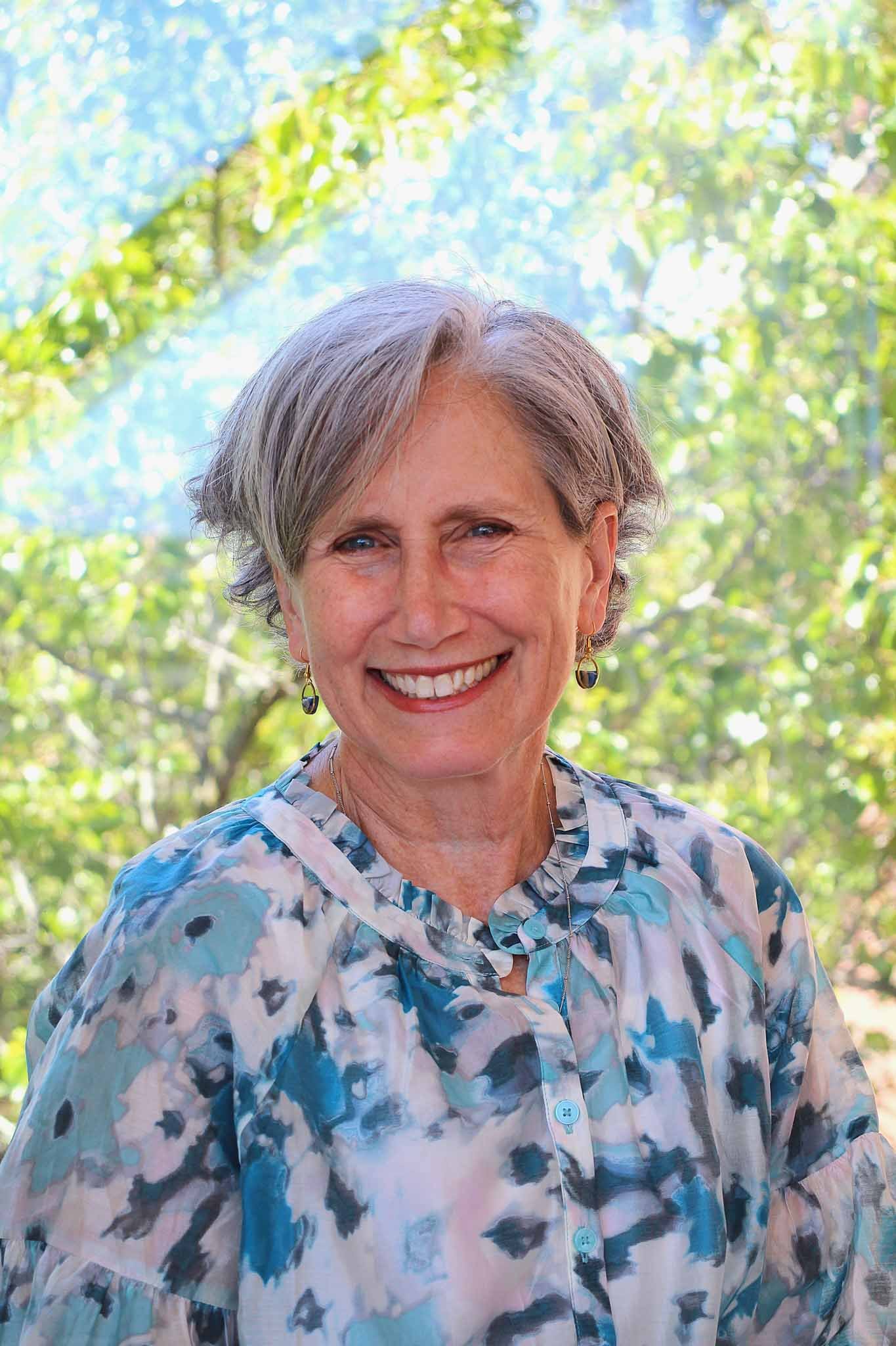 A smiling woman with gray hair wearing a blue polka dot top outdoors in front of green trees.
