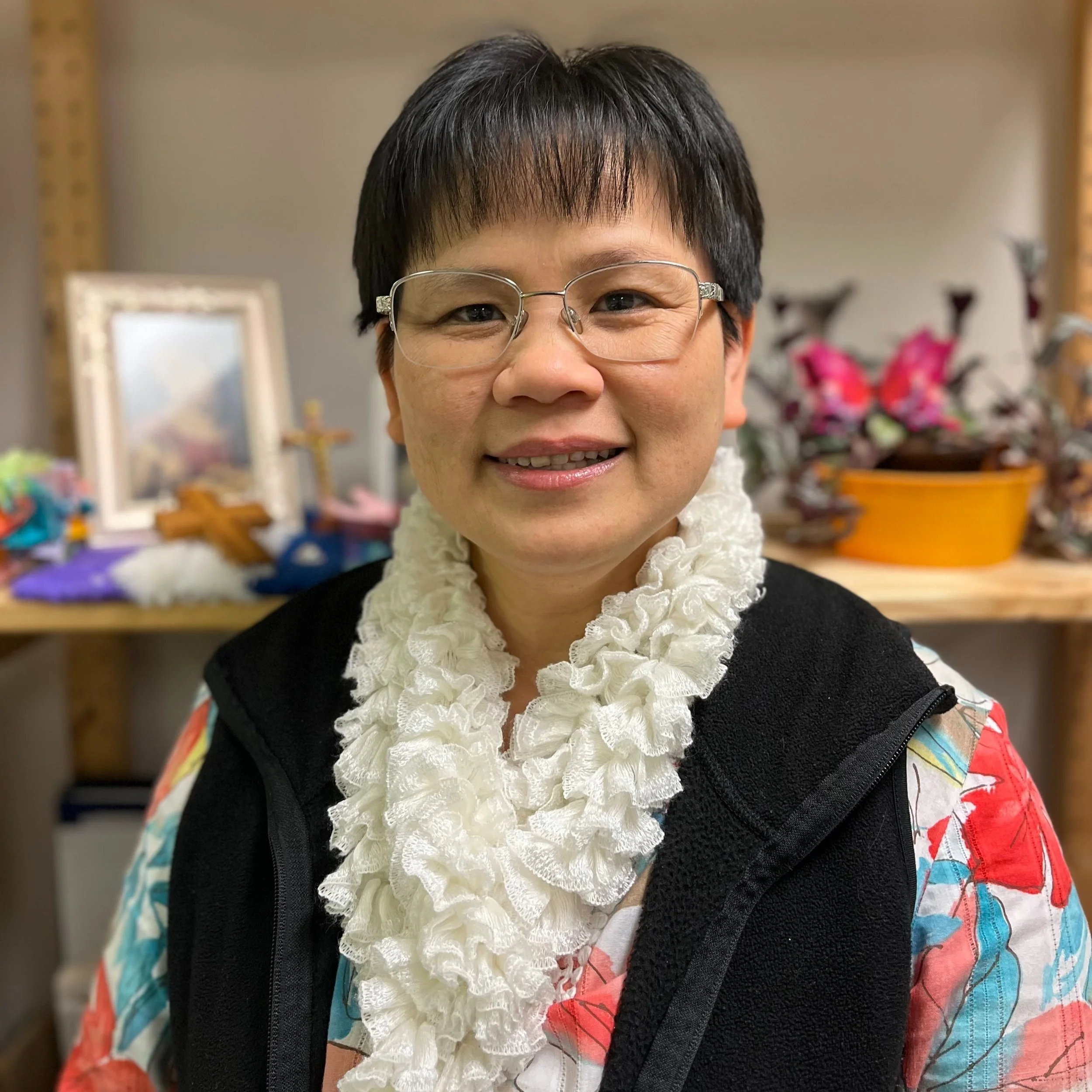 Person wearing glasses and a white scarf, standing indoors with a shelf and potted plant in the background.
