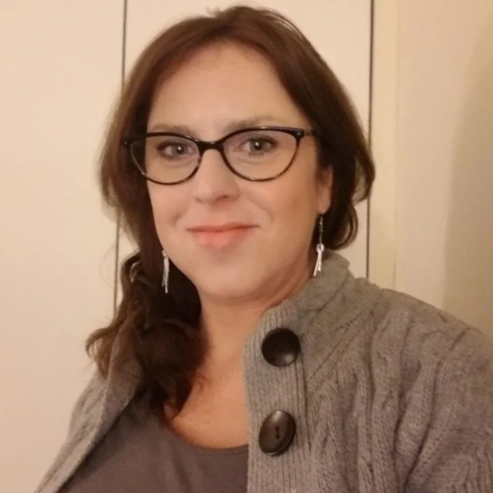 Woman with glasses and brown hair wearing a gray sweater and earrings, smiling indoors.