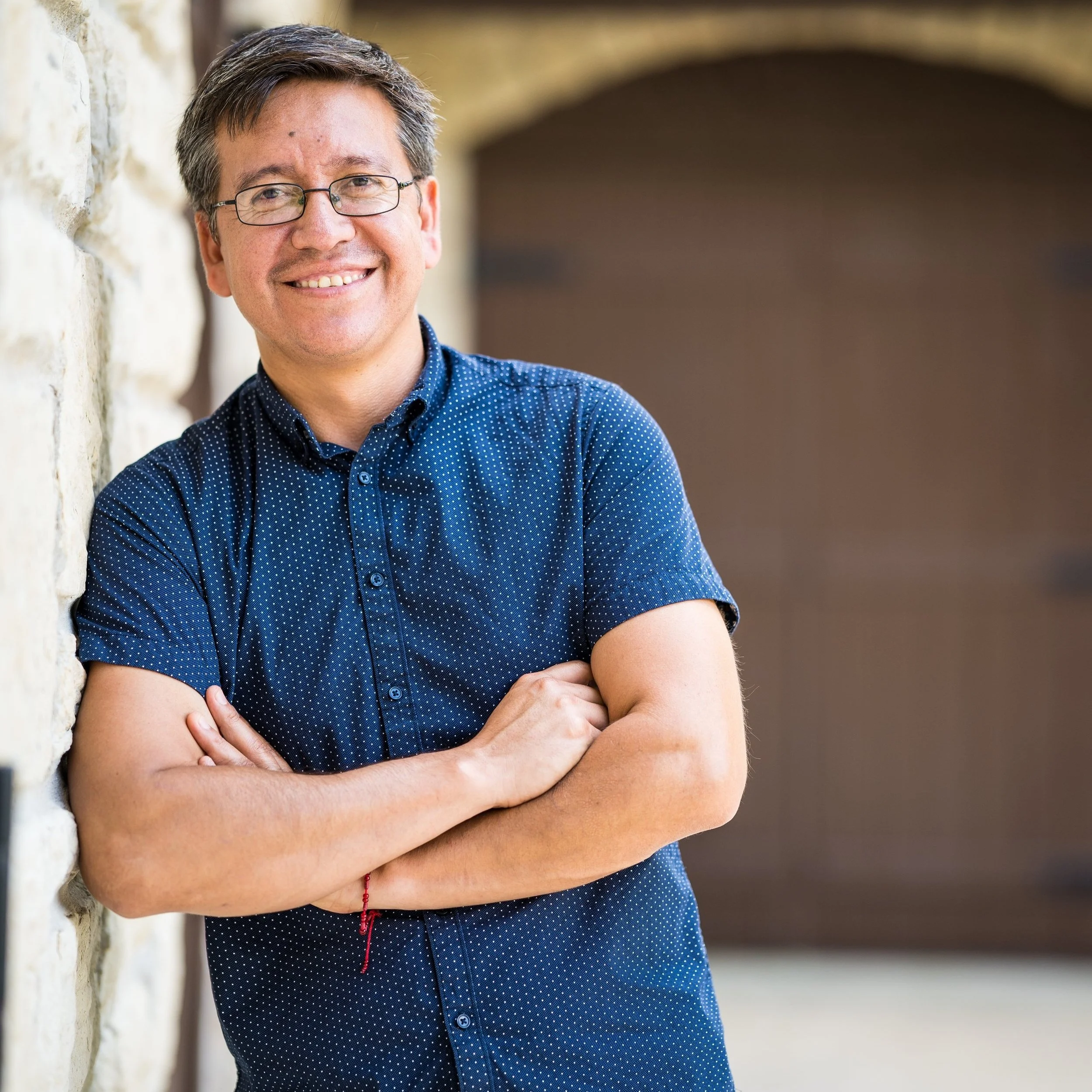 Man in blue dotted shirt leaning against a stone wall, smiling with arms crossed, wearing glasses.