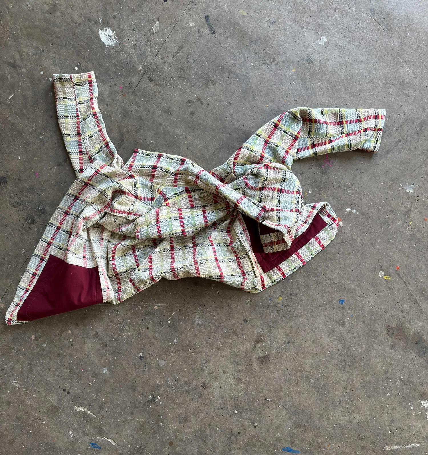 A chequered cream and pale blue coat with deep red lining lies with arms open on a concrete floor - an artwork from Andi Walker's solo exhibition