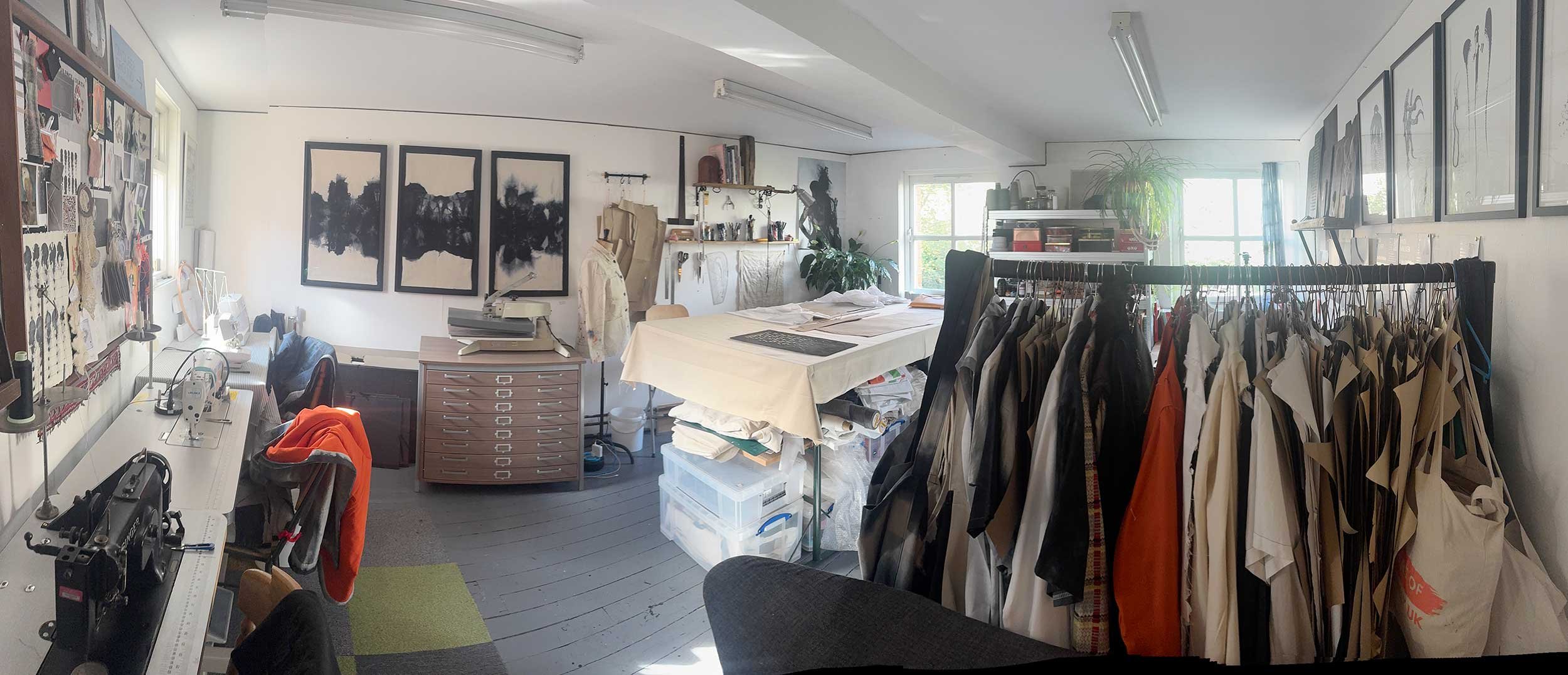 A wide angled photograph of Andi Walkers studio - showing busy table, tools, a sewing machines, clothing on a rail, ink and other art on walls,