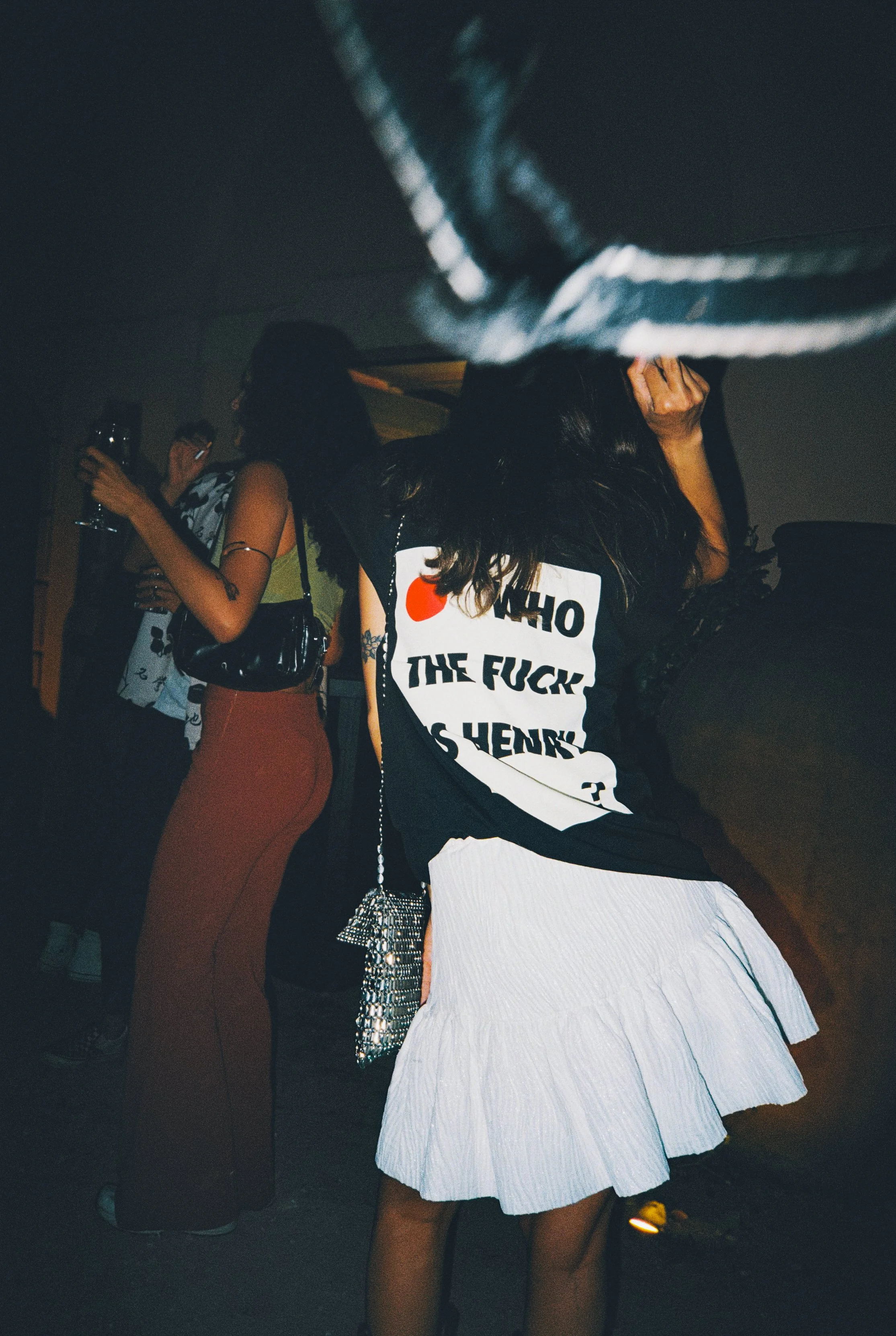 People at a party, one person wearing a black shirt with bold text and a white ruffled skirt, another person in brown pants and a sleeveless top, some holding drinks, in a dimly lit indoor setting.