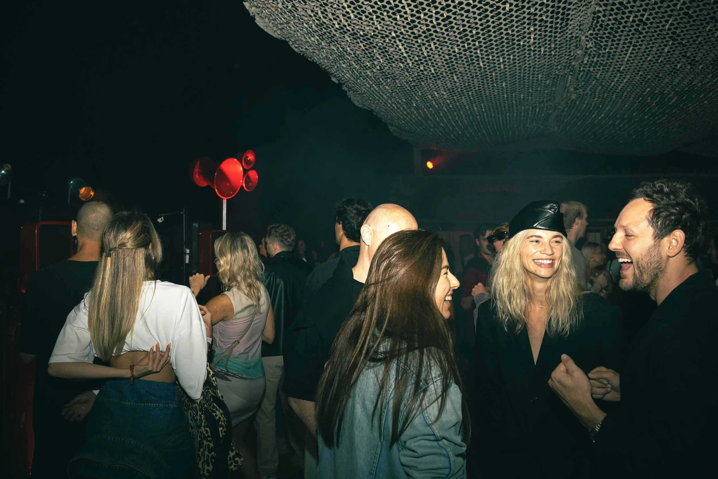 People socializing and laughing at a nightclub or party with dark lighting and red speakers in the background.