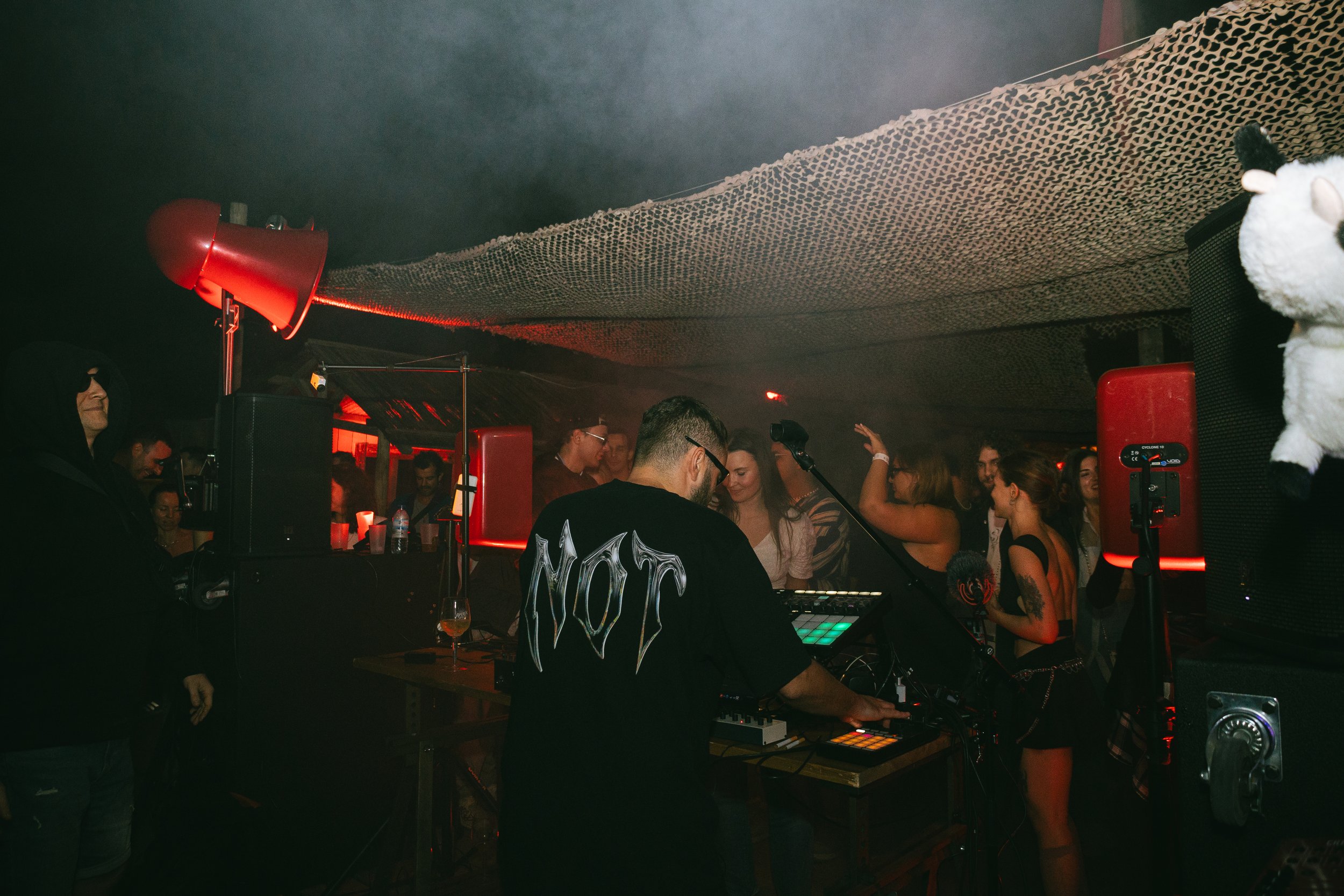 A DJ performing at a crowded party with people dancing, red lighting, and a dark ambiance.