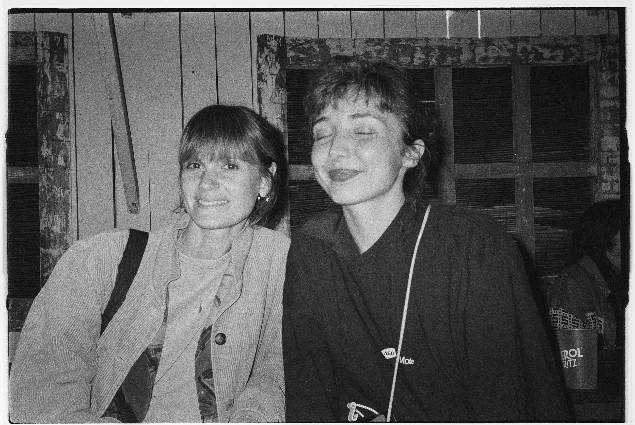 Two women sitting close together, one smiling with eyes open, the other with eyes closed and smiling, in a rustic indoor setting with wooden walls.