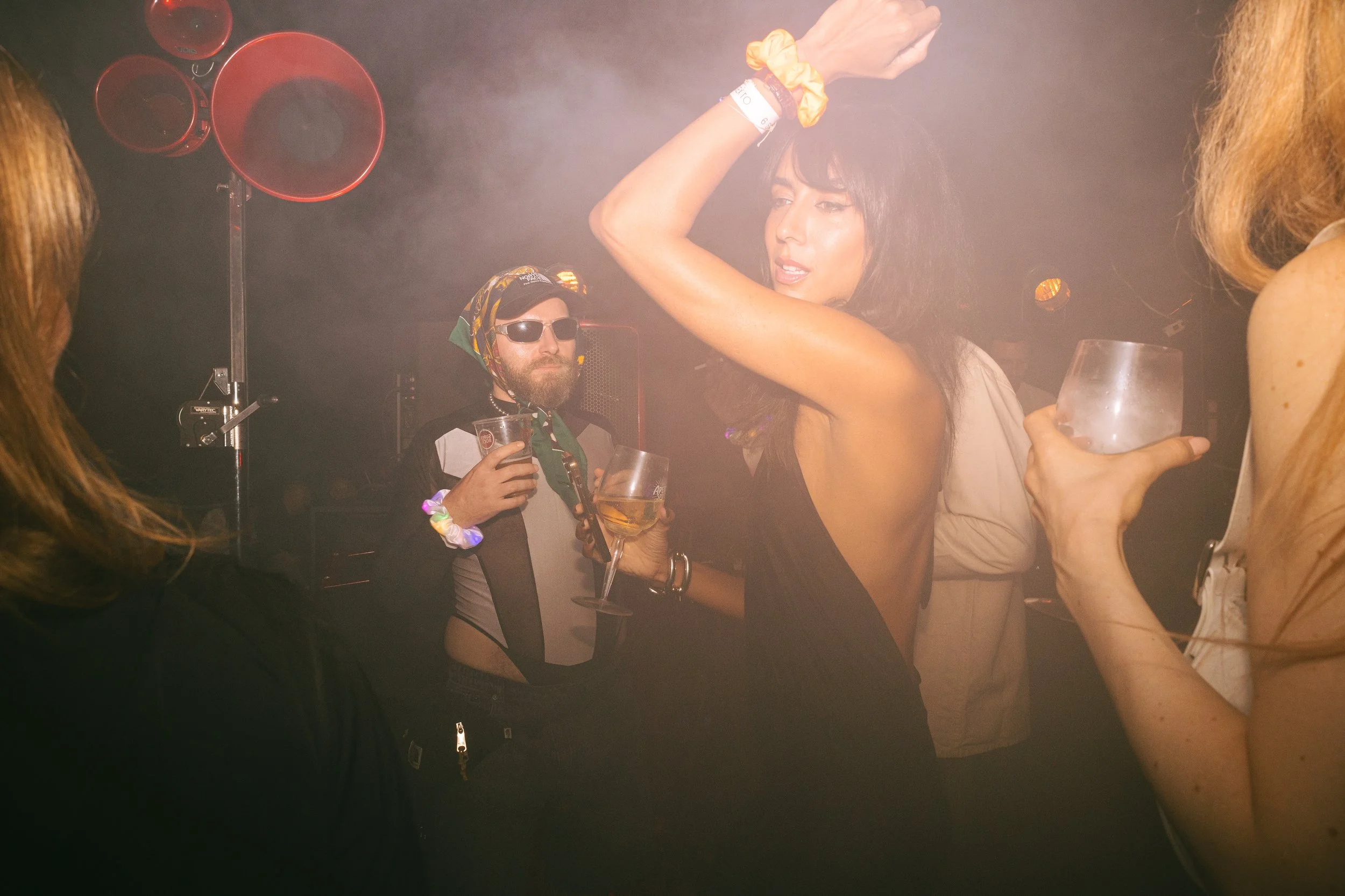 People dancing and socializing at a nightclub, with a woman in a black dress dancing in the foreground and a man in sunglasses holding a drink in the background.