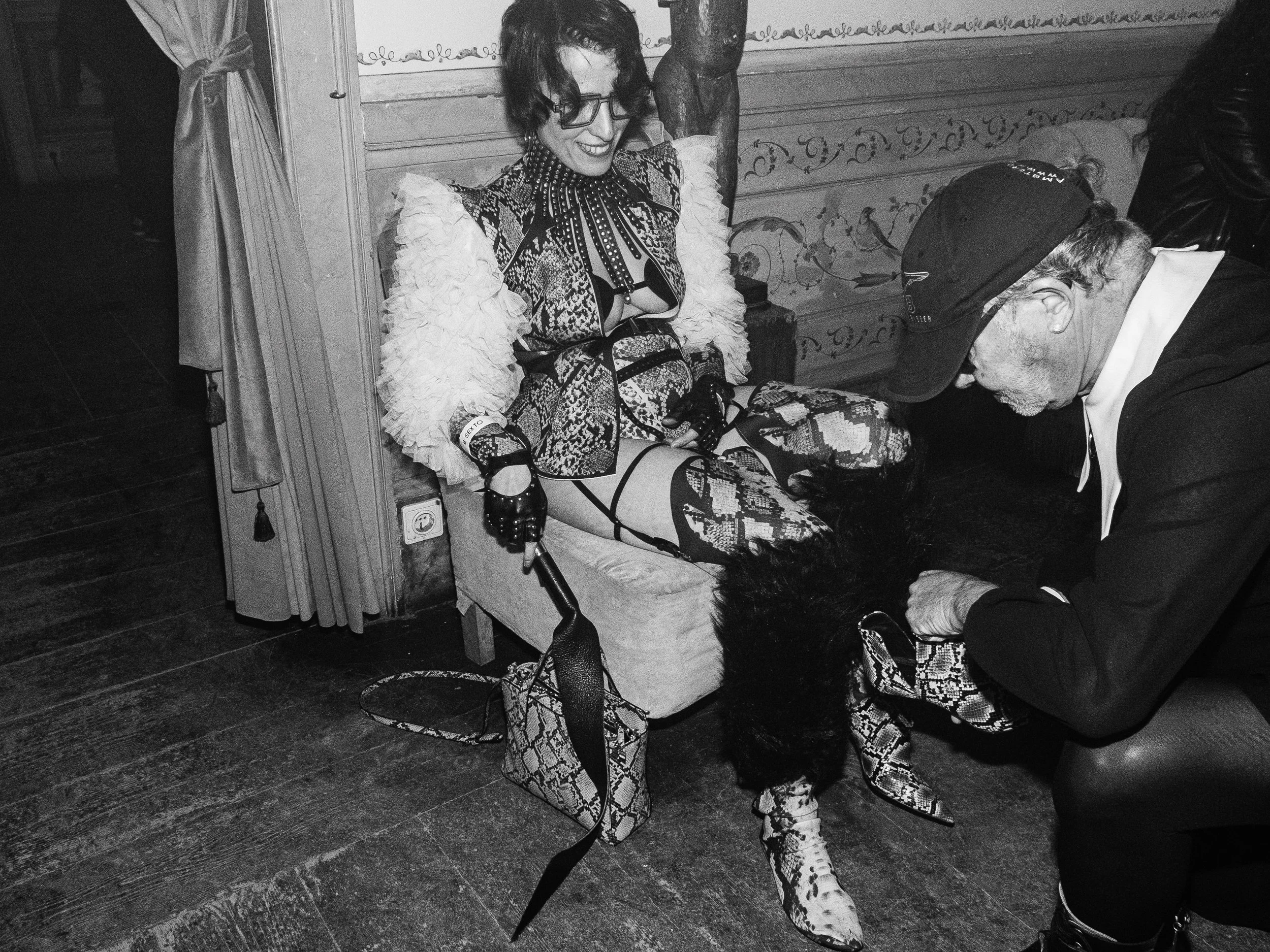 A woman sitting on a cushioned chair in a fashionable outfit, with a man leaning over her, adjusting her shoe. The woman is wearing patterned clothing and high-heeled snakeskin boots, while the man is dressed in dark clothes and a baseball cap. The s