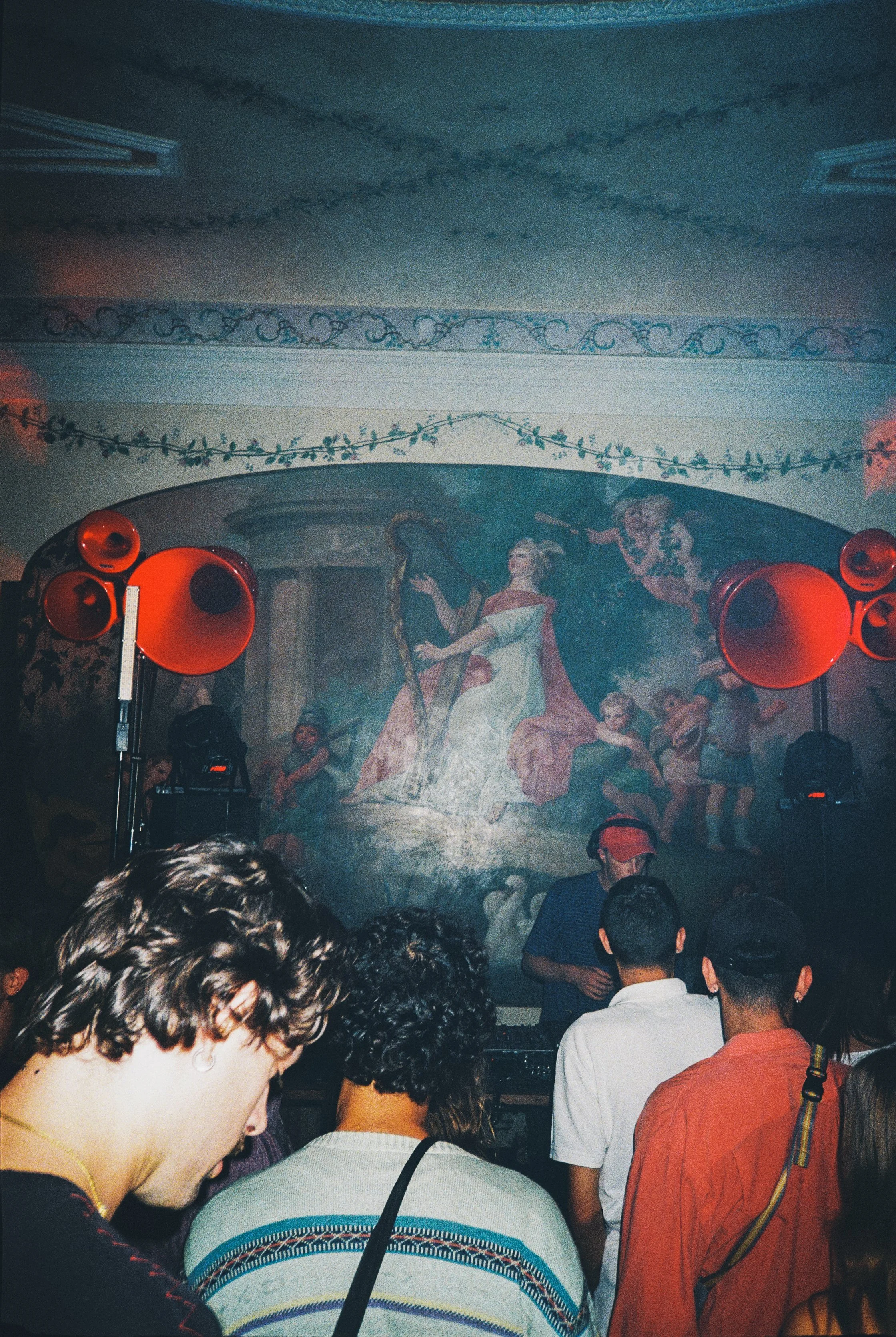 A crowded indoor event with a mural painting in the background and red loudspeakers on stands, with people standing and listening.