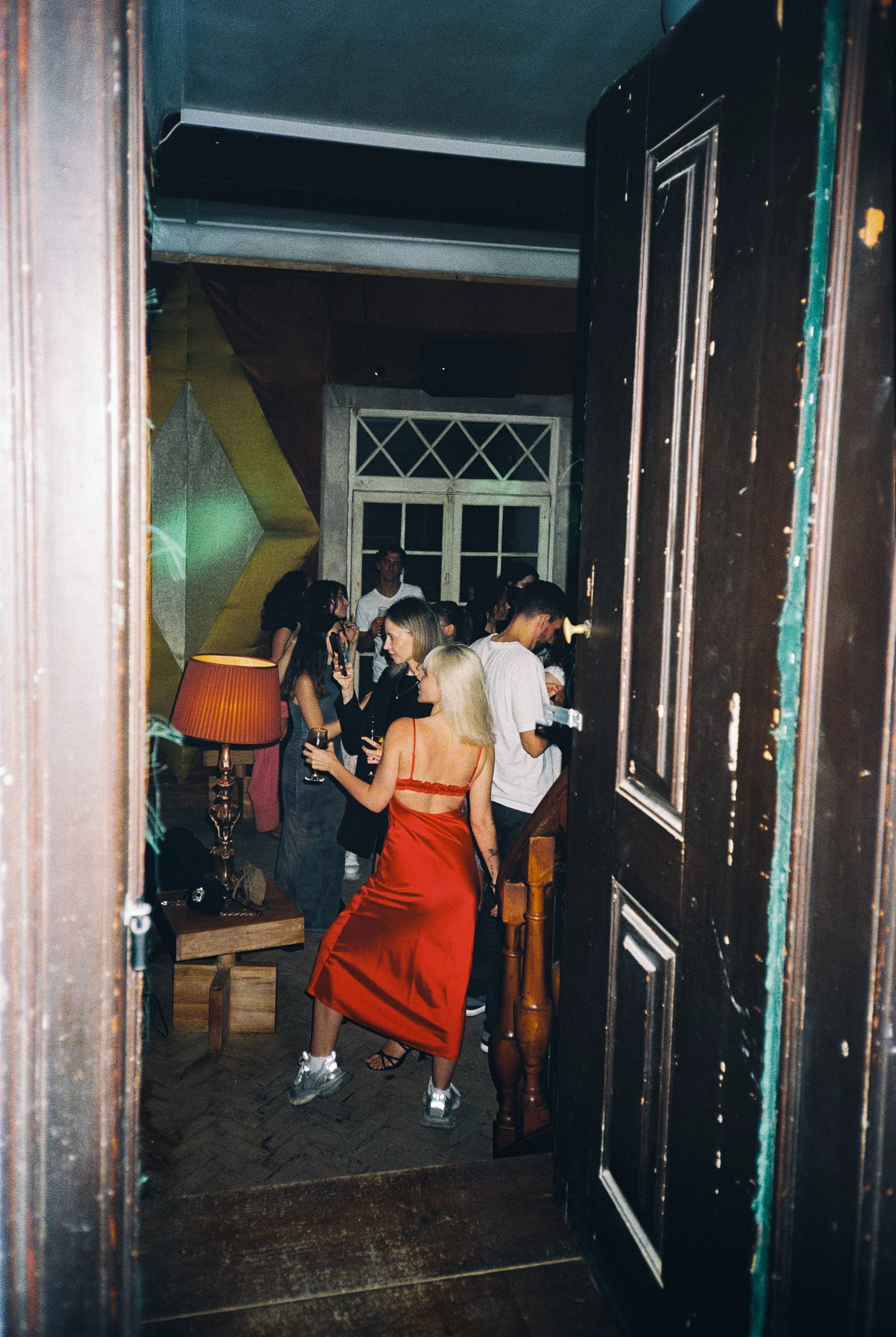 People socializing at a party in a dimly lit room, some holding drinks, with a woman in a red dress in the foreground.
