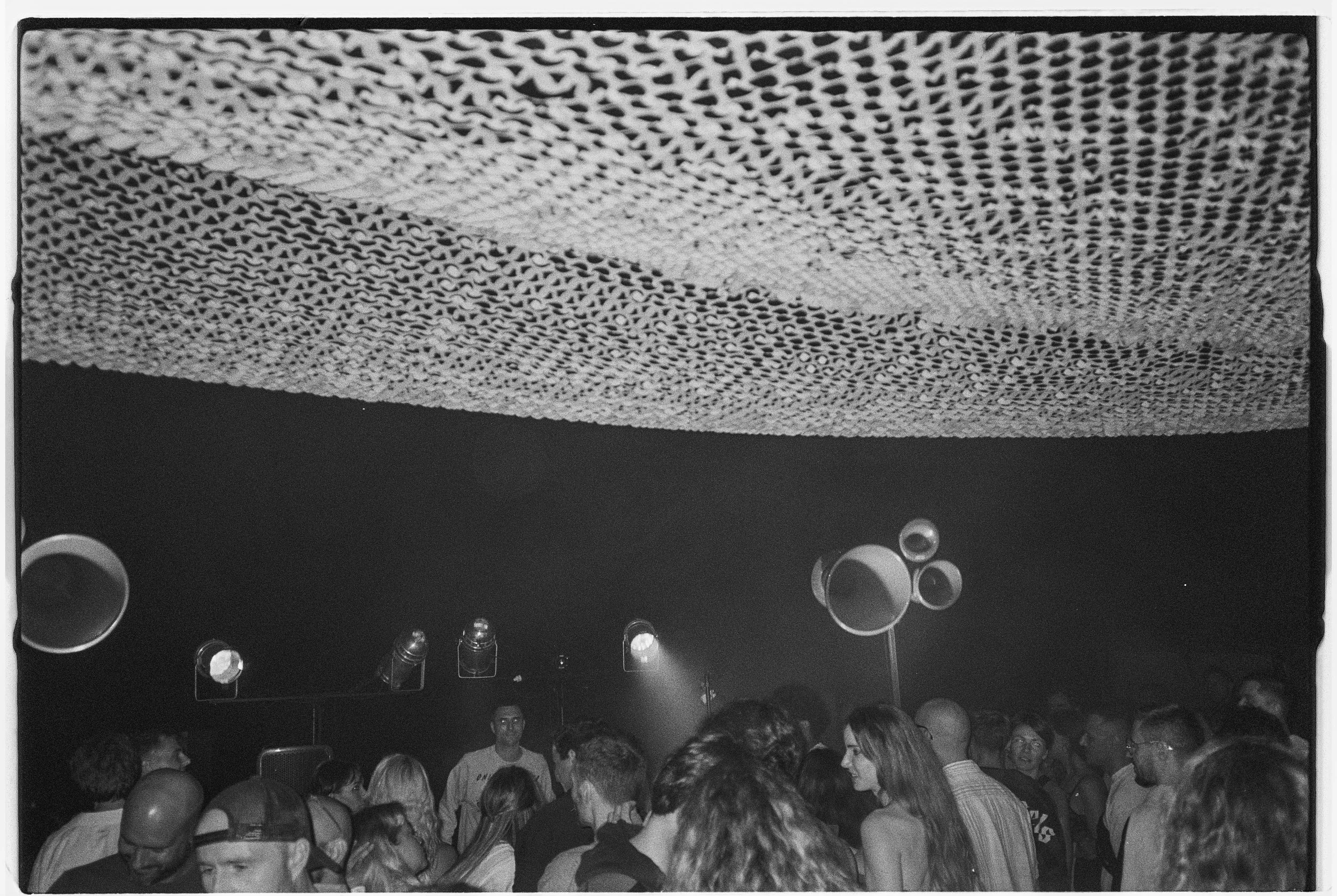 Black and white photograph of a crowded dance club with a textured ceiling, multiple large speakers and a person at the DJ booth in the background.