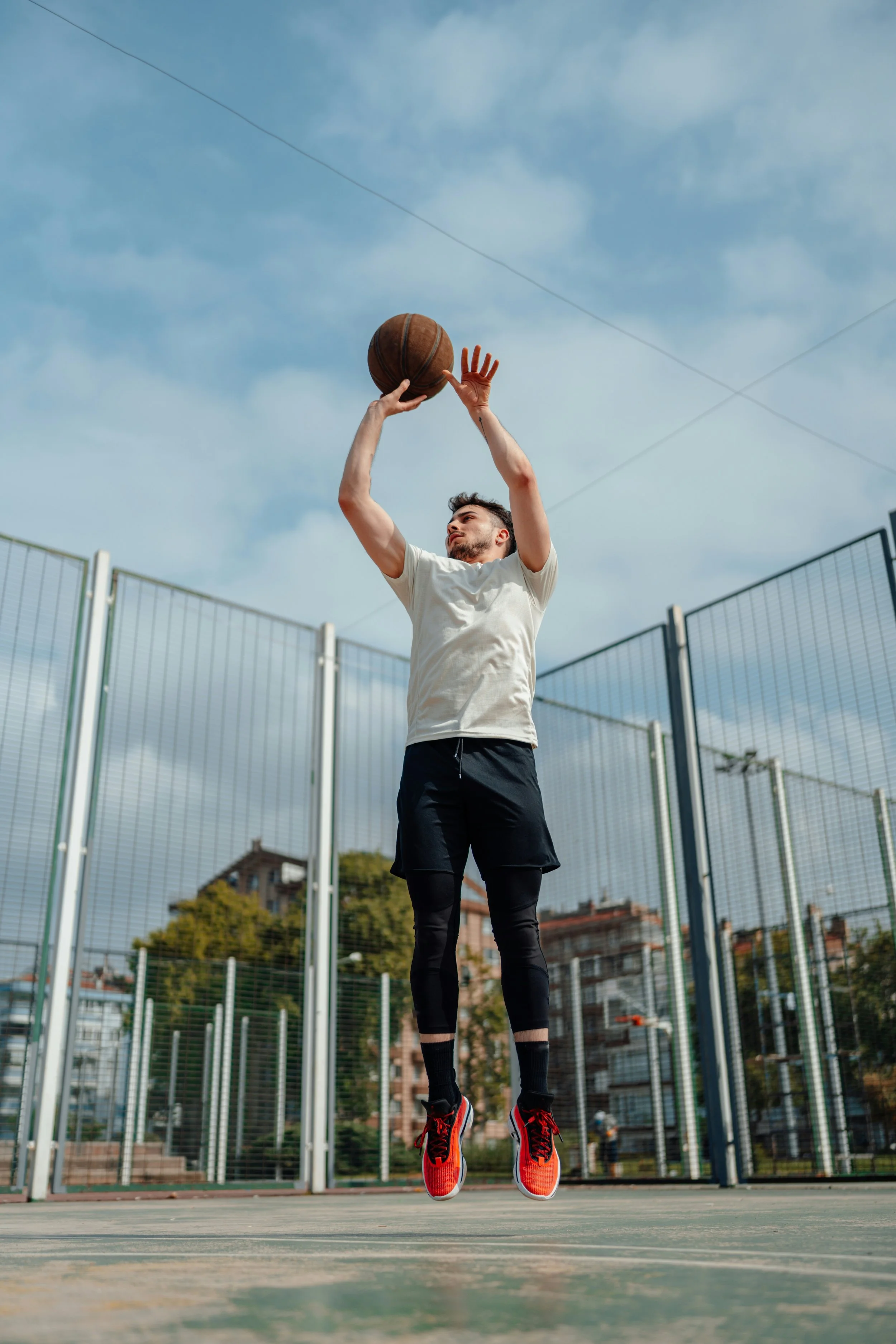 Man playing basketball, jumping and shooting, wearing a white shirt and red sneakers, outdoor court.