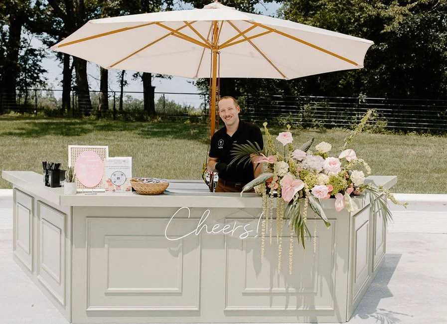 Man standing behind an outdoor bar decorated with a large floral arrangement, with a white umbrella overhead, on a sunny day.