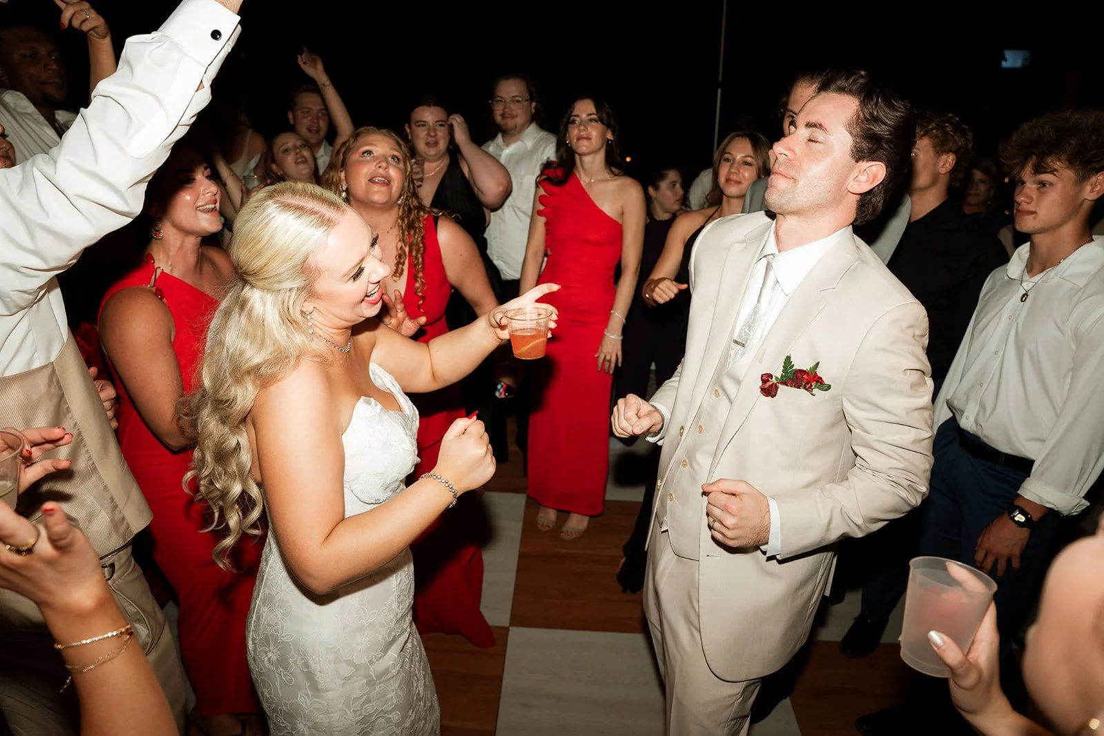 A group of people dancing and celebrating at a wedding reception, with a bride in a white dress and a groom in a light suit at the center.