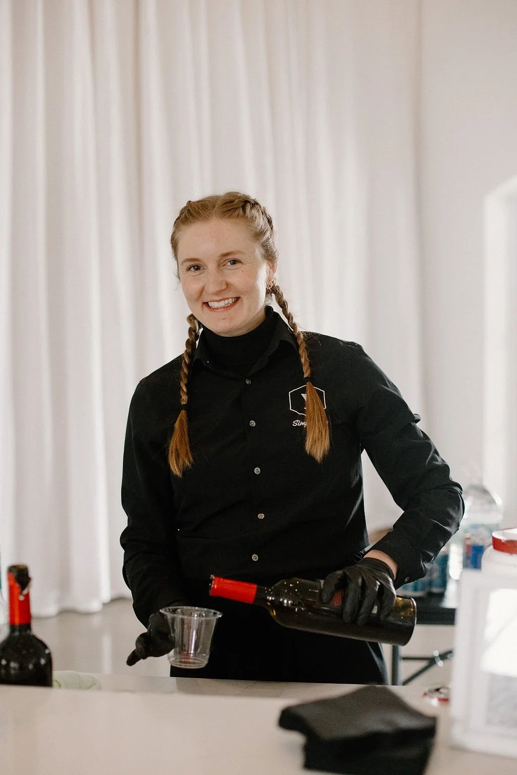 A woman wearing a black shirt, black gloves, and a black turtleneck pouring wine into a glass, smiling at the camera, with a white curtain background.