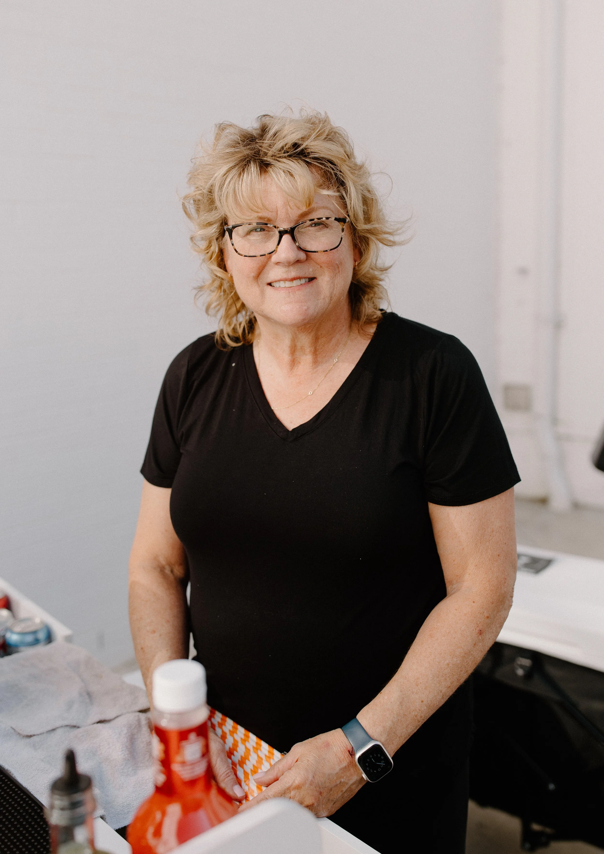 A woman with curly blonde hair, glasses, and wearing a black shirt, working at a table with medical supplies and bottles, smiling at the camera.